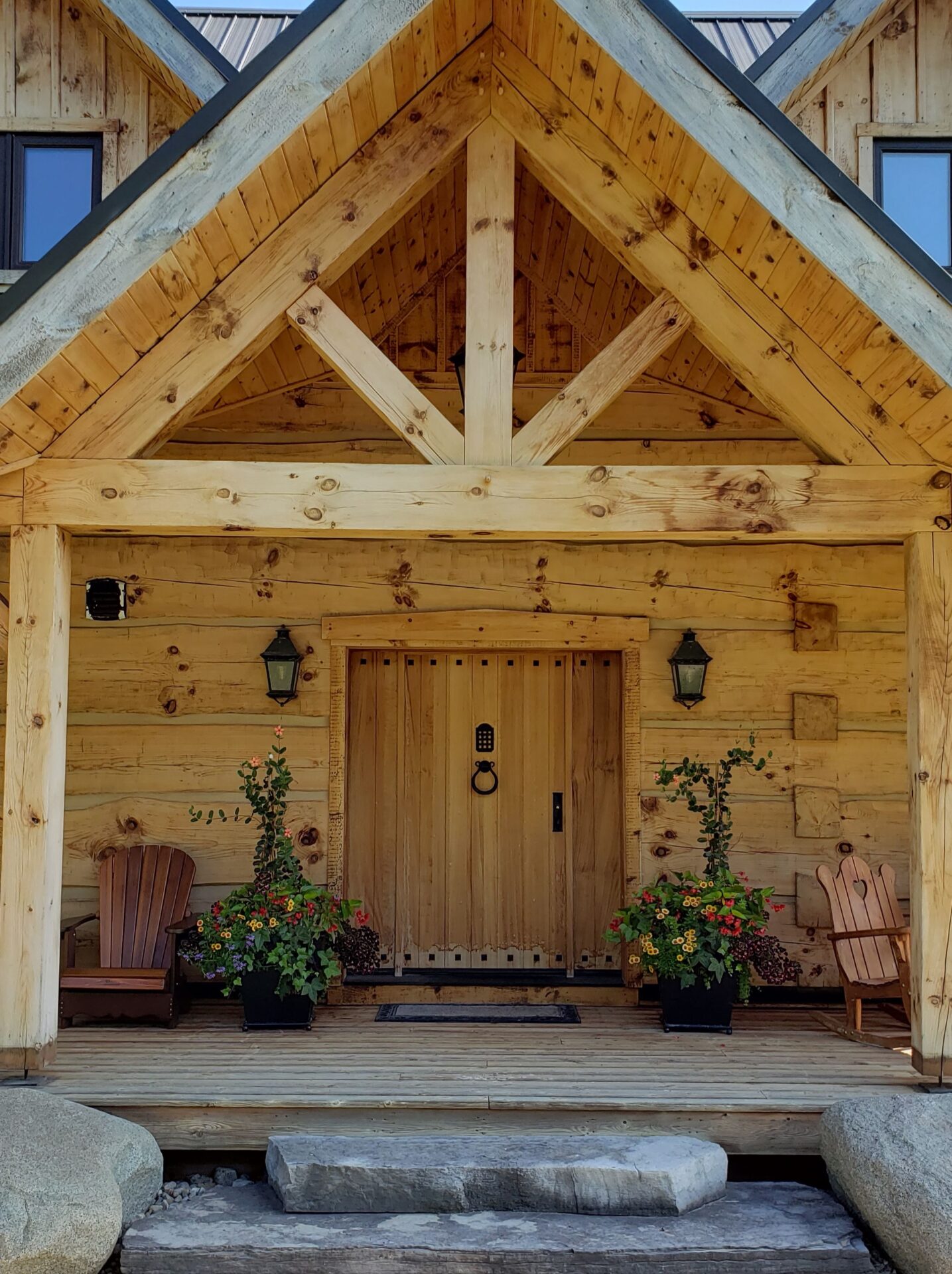 The image shows a wooden cabin entrance featuring a large door adorned with metal hardware, flanked by Adirondack chairs and vibrant potted flowers.