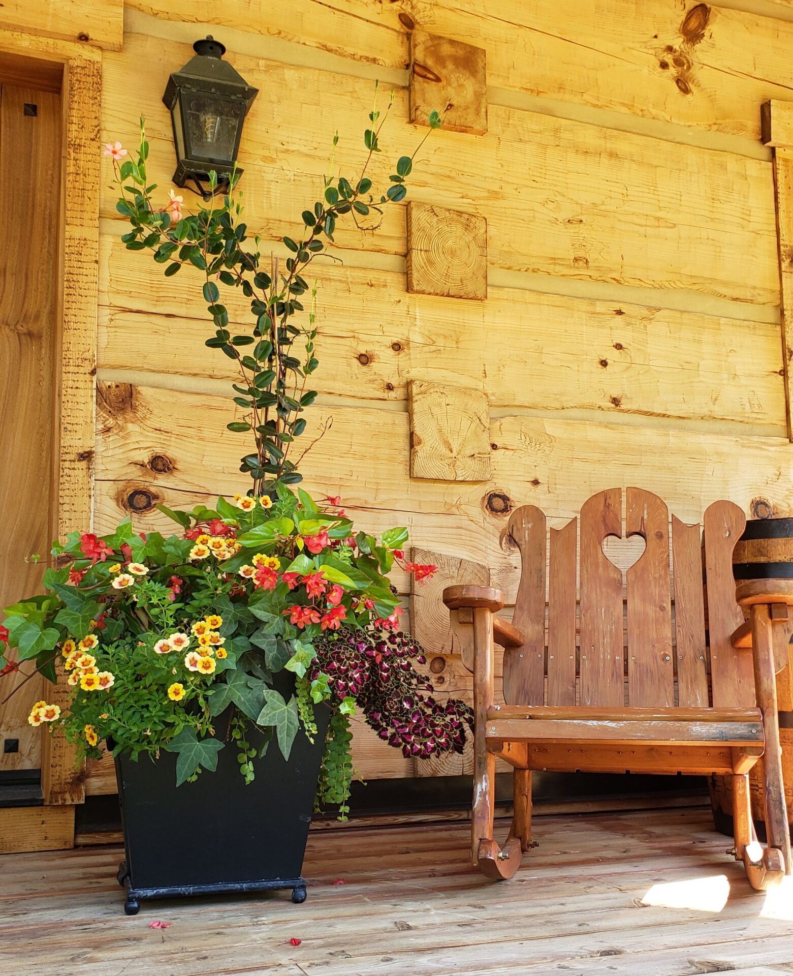 A wooden porch with a rustic bench featuring a heart design, a vibrant potted plant arrangement, and a classic lantern fixture on a sunny day.