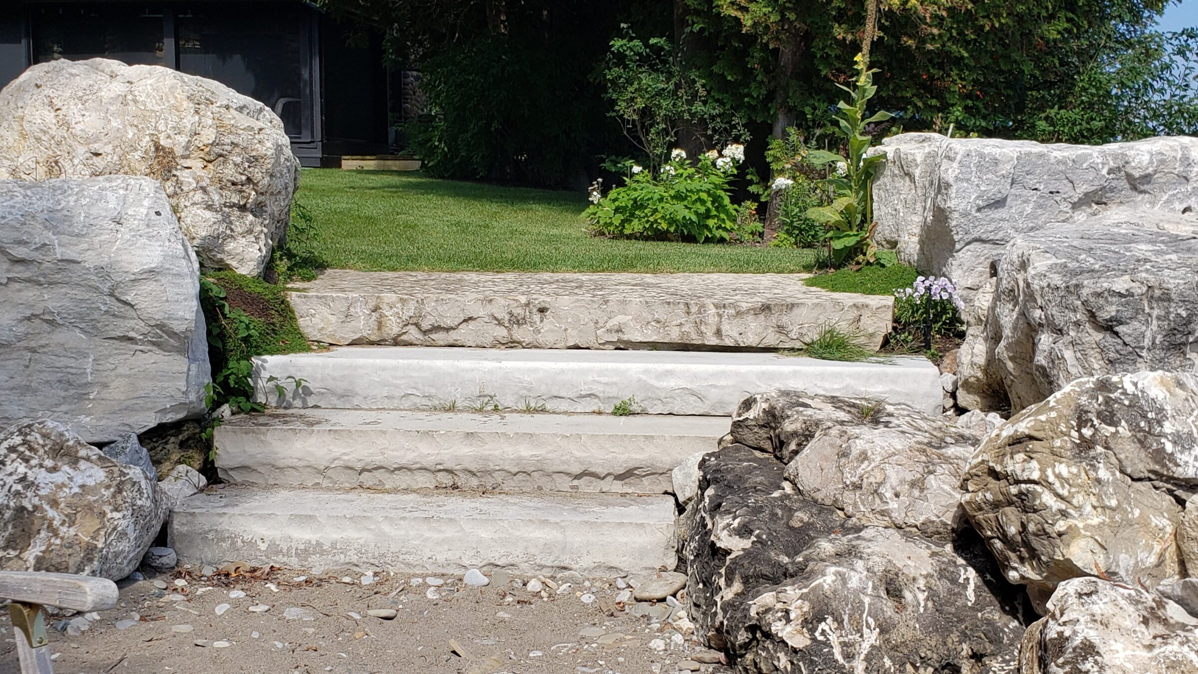 Stone steps flanked by large boulders lead up to a green lawn with flowering shrubs. A sunny day casts shadows on the serene garden.
