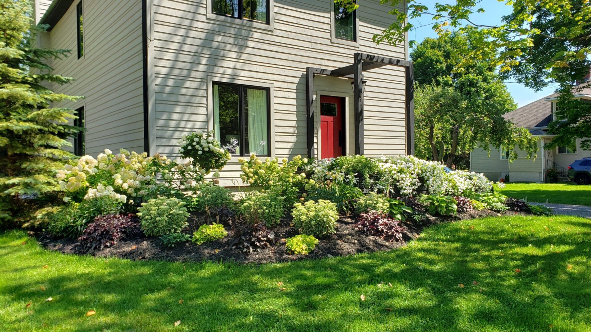 A beige house with green shutters, red door, and a front garden filled with hydrangeas and other greenery under a sunny sky.