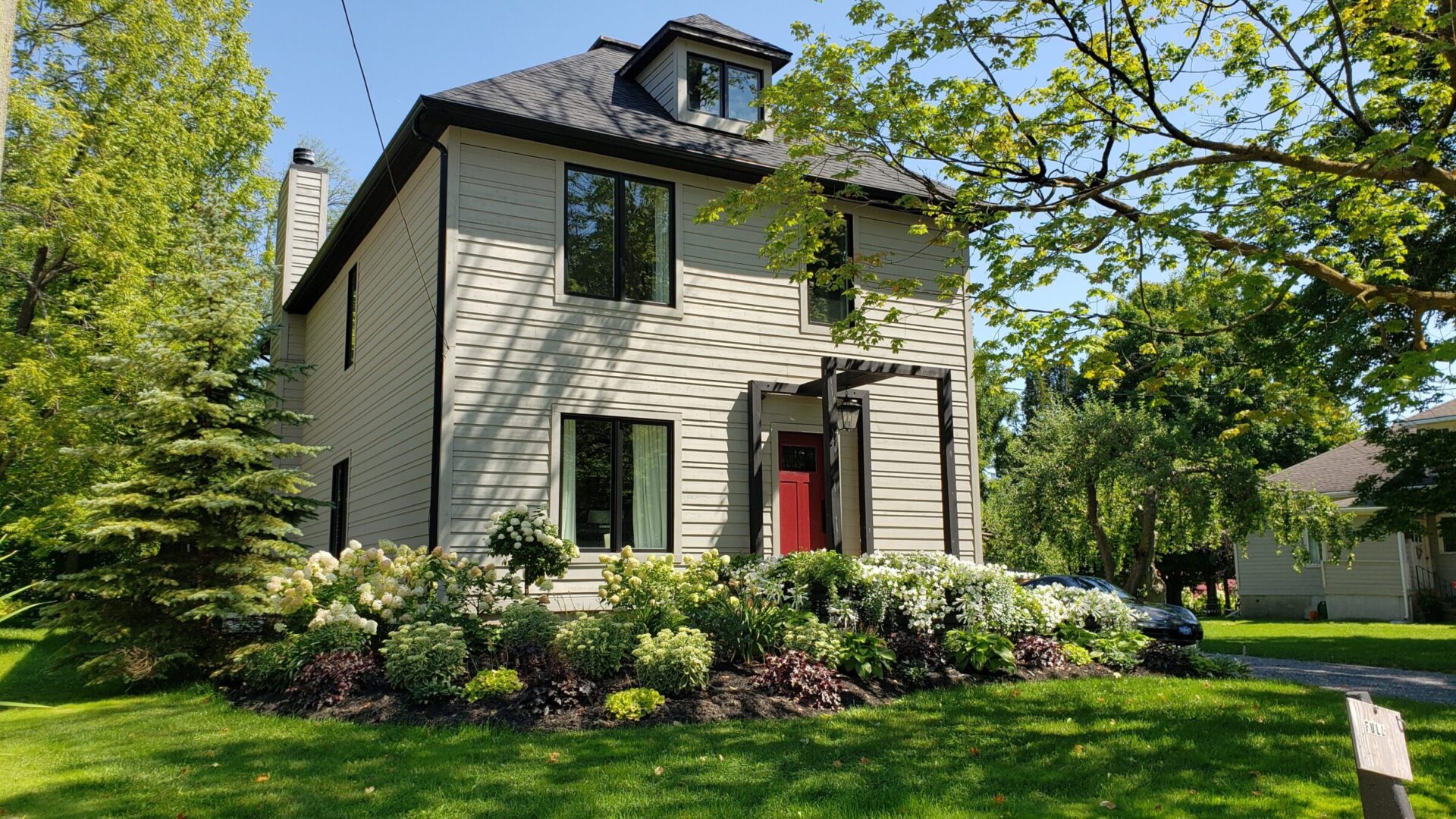 A two-story house with beige siding and a red door surrounded by lush greenery and hydrangea bushes under a clear blue sky.