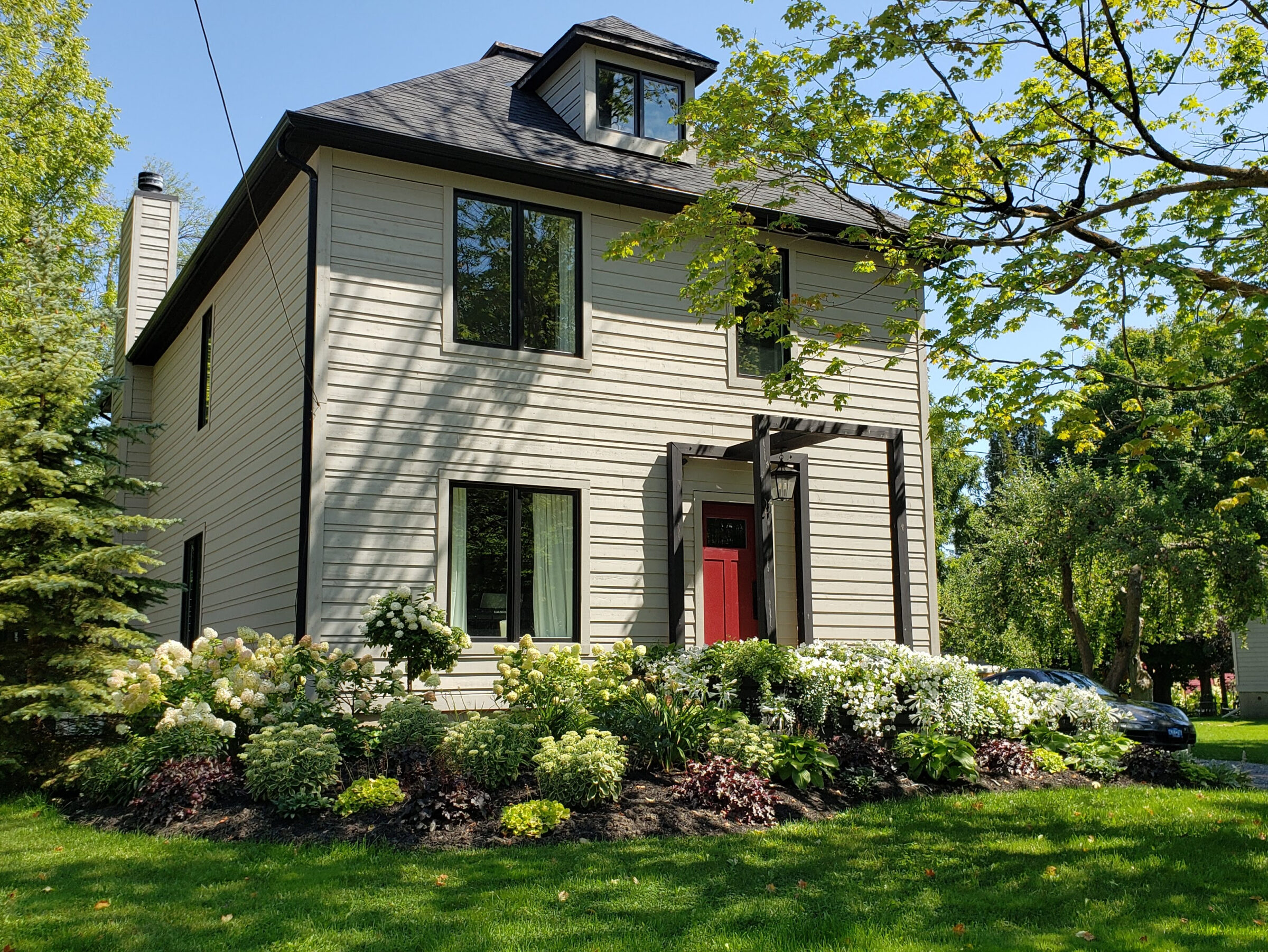 A two-story home with beige siding and a red door is surrounded by lush gardens, with a car parked by the curb, on a sunny day.