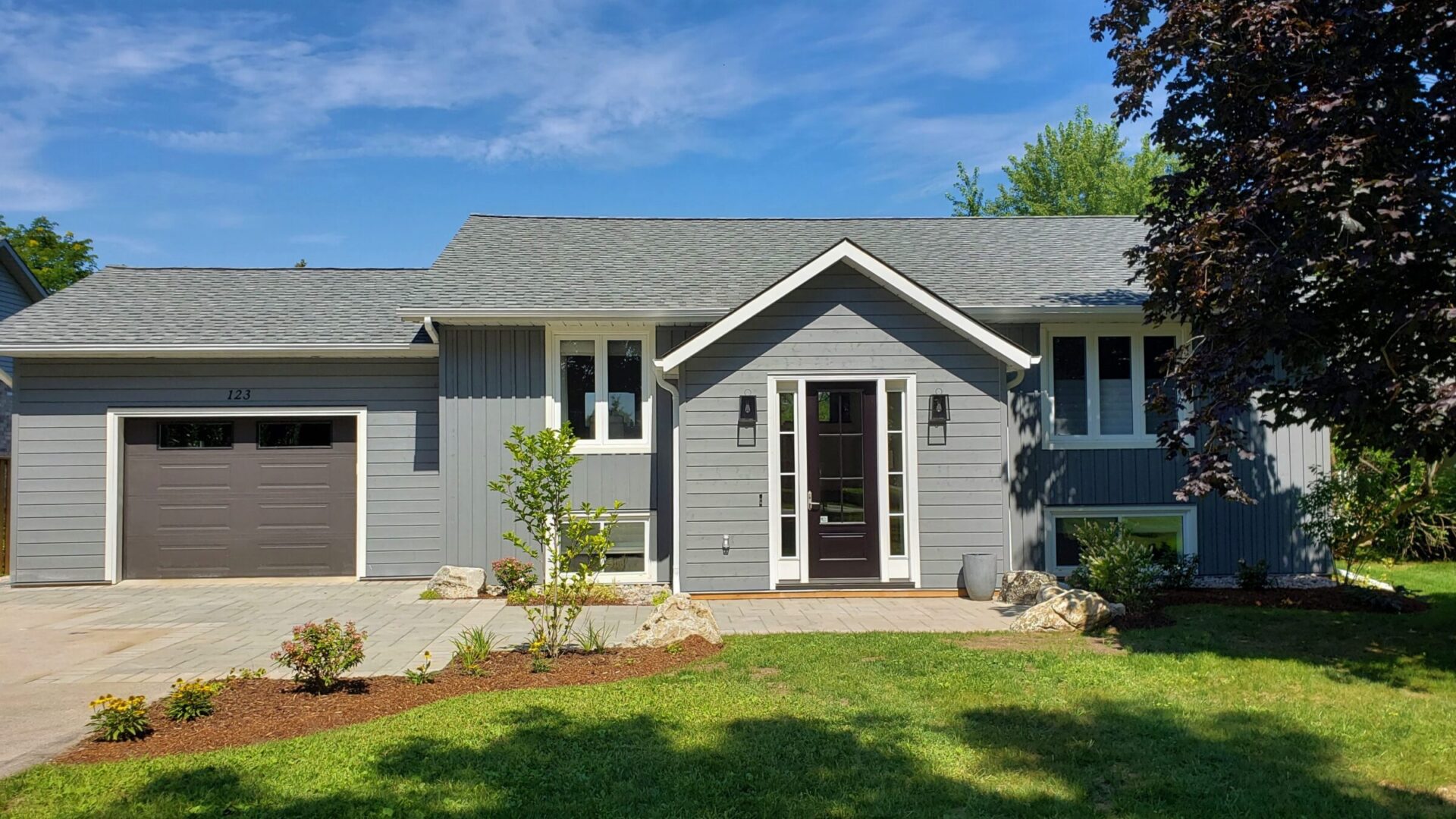 A single-story gray house with white trim, a garage, and a landscaped front yard under a clear blue sky. Trees and shrubs are partially visible.