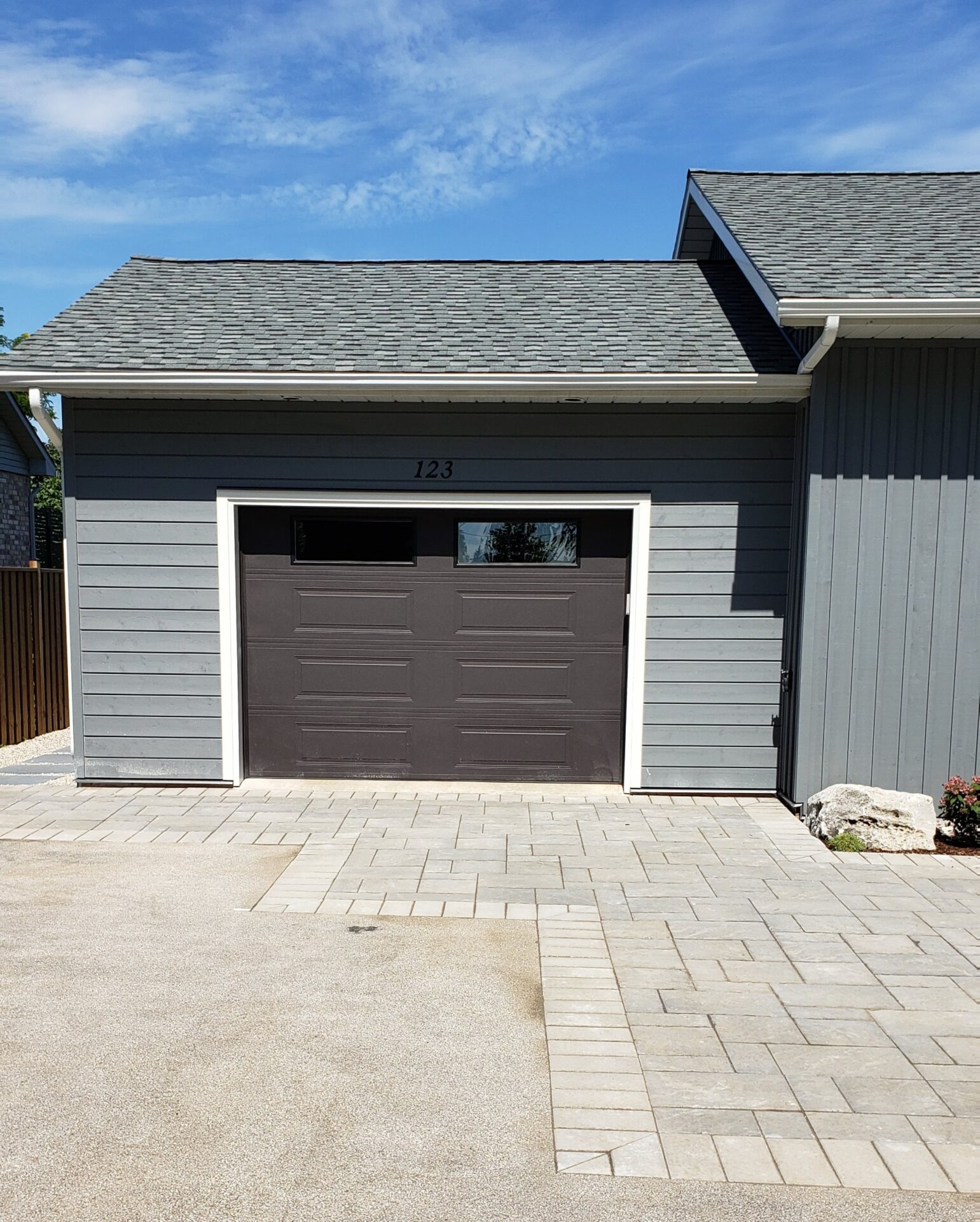 This image shows a grey detached garage with a dark garage door featuring small windows and house number 