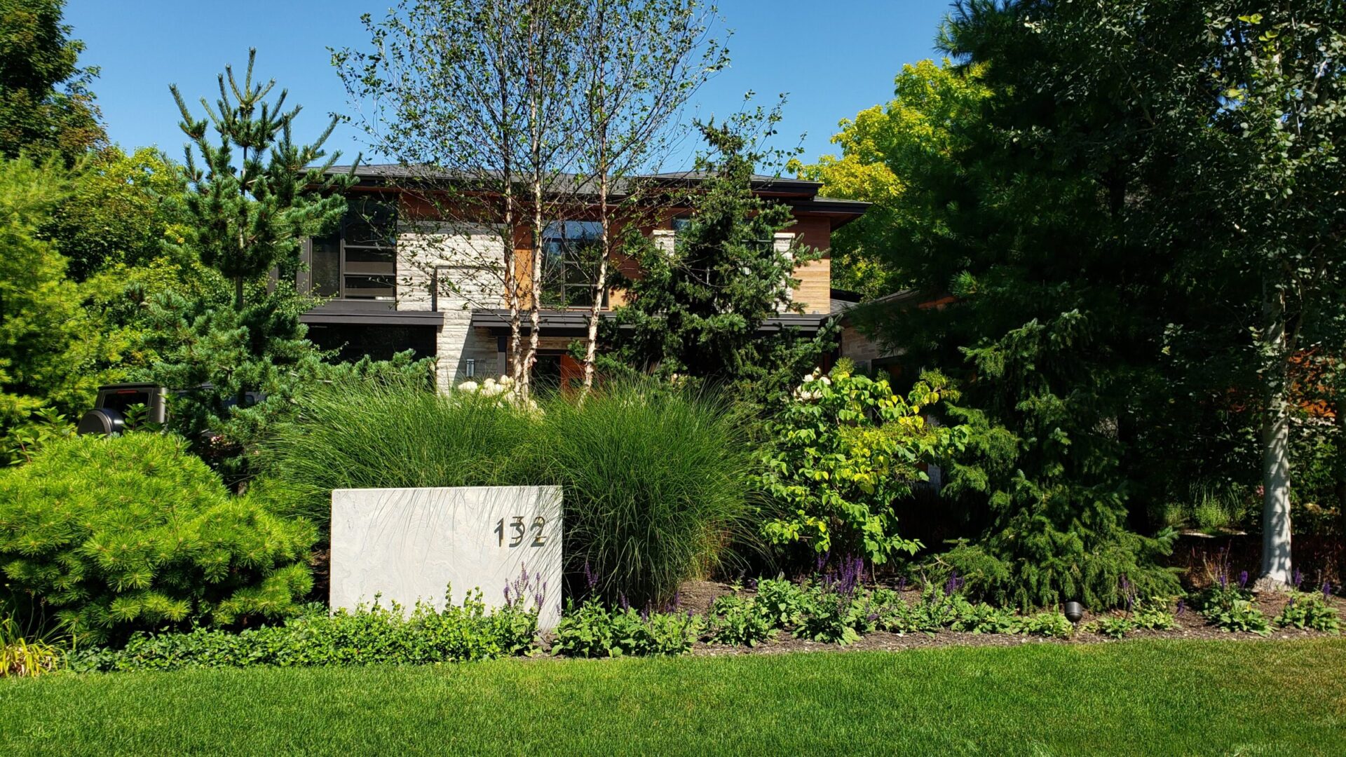 A well-maintained garden in front of a modern house with lush greenery, a variety of plants, and a prominent address stone marked 