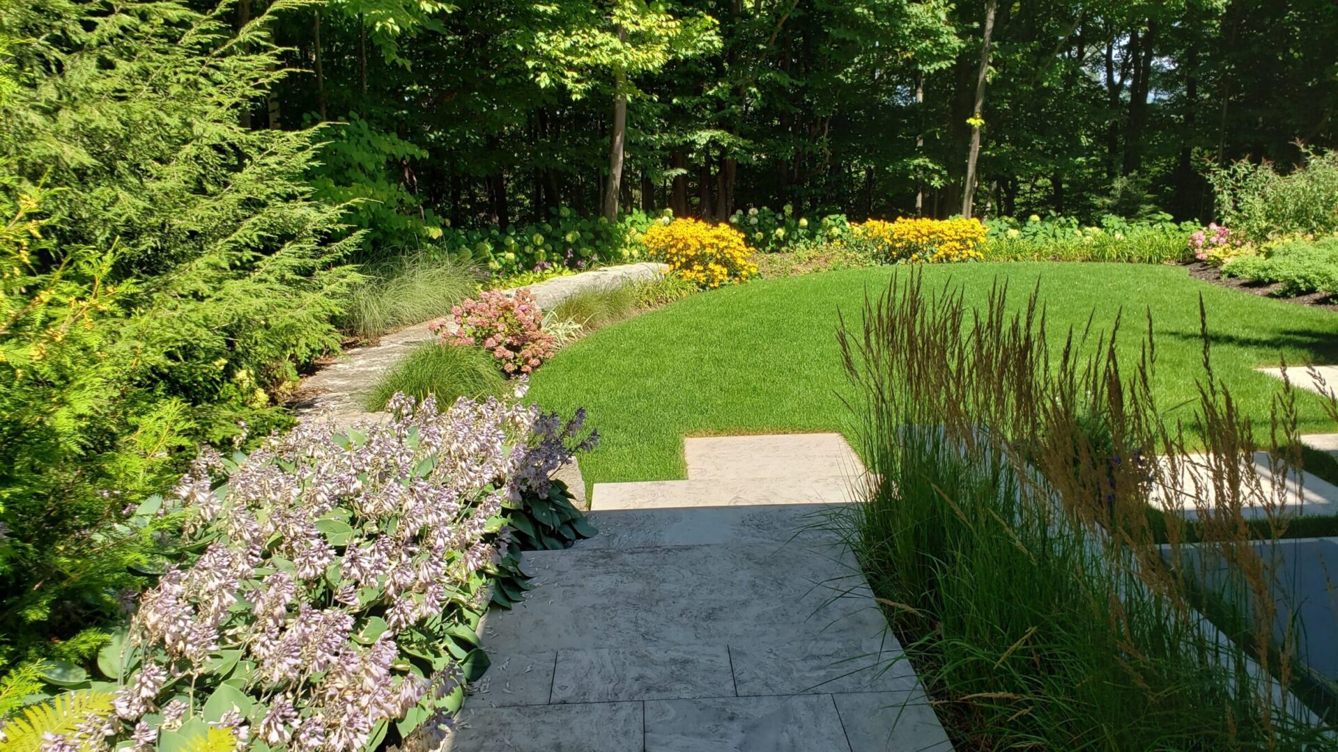 A well-manicured garden with diverse plants, a lush lawn, stone path, flowering shrubs, and a backdrop of green trees under a clear blue sky.
