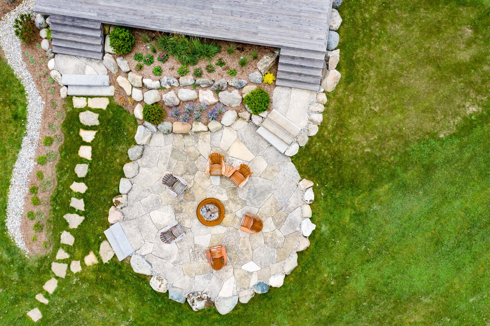 Aerial view of a landscaped outdoor area with a stone patio, fire pit, wooden chairs, surrounded by green grass and a wooden structure.
