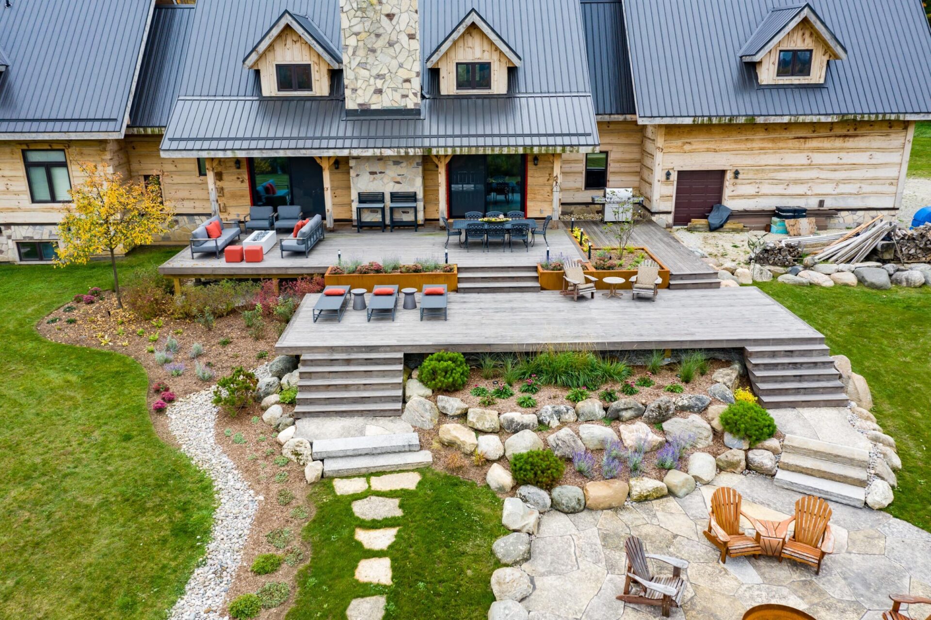 This image shows a rustic log cabin with a modern outdoor terrace, a stone fire pit area, lush landscaping, and cozy patio furniture.