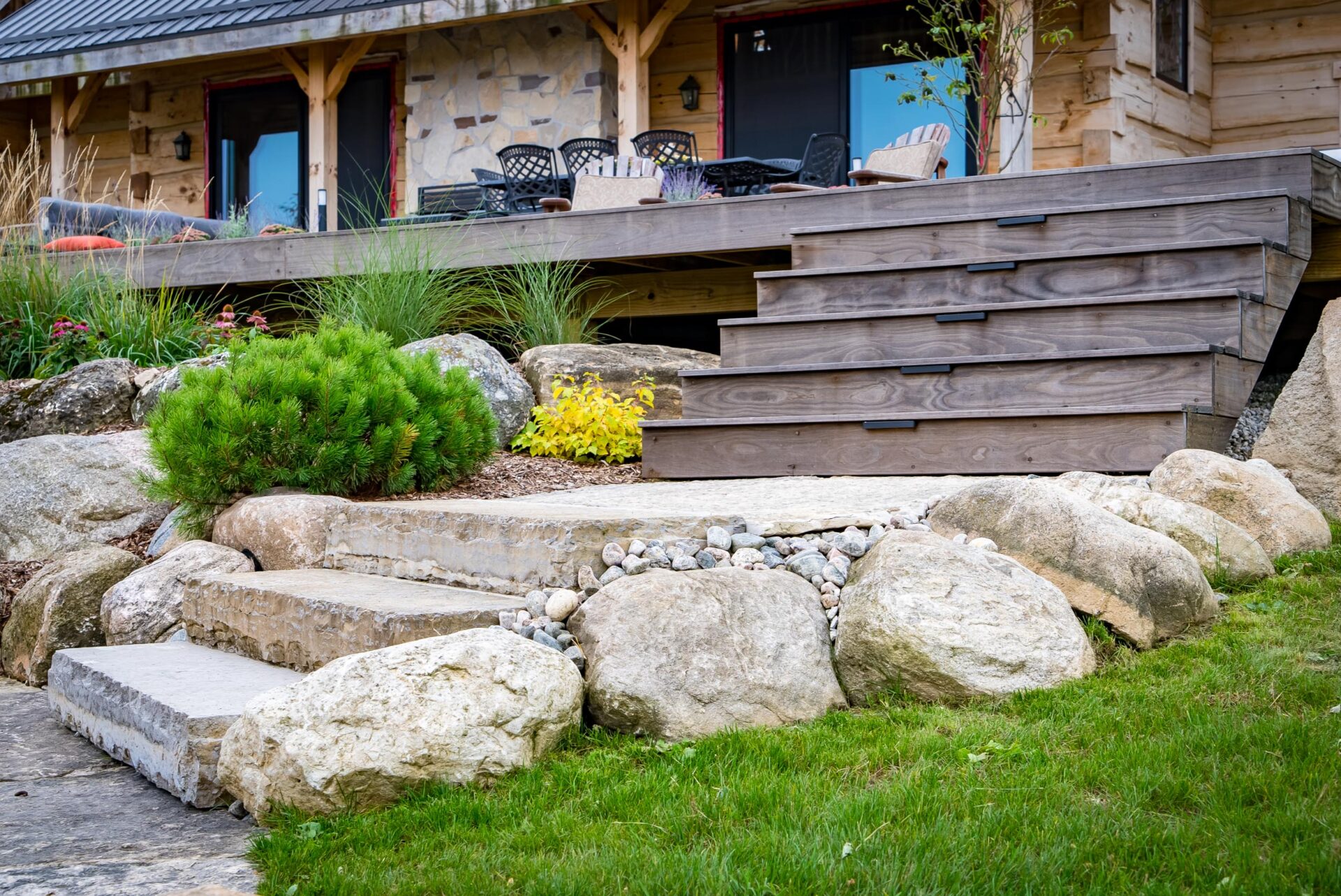A rustic wooden house with a deck, landscaped with natural stone steps, boulders, and green plants. Outdoor furniture is visible in the background.