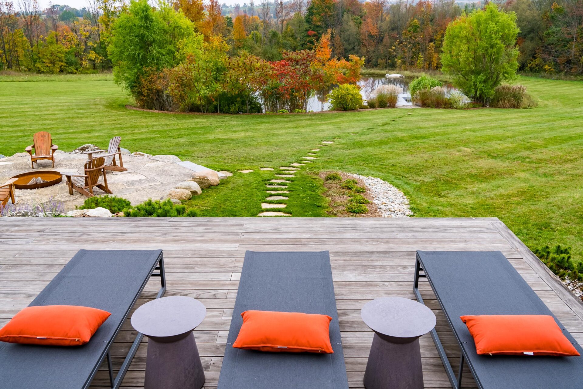 A serene outdoor setting with lounge chairs, orange cushions, a fire pit with Adirondack chairs, and a lush landscape with a pond in autumn.