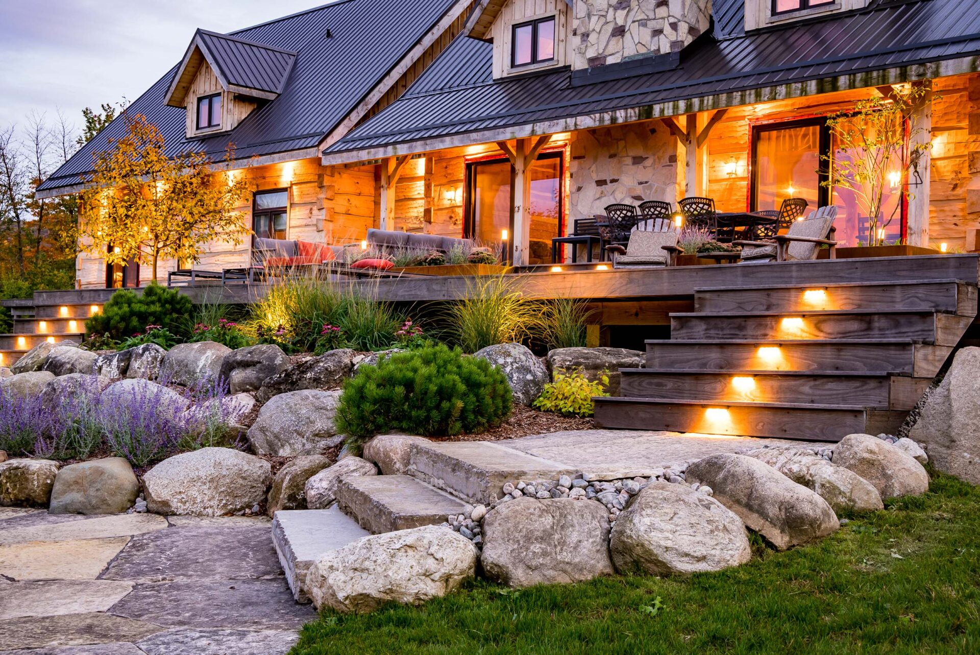 A rustic house with a lit porch, wooden steps with lights, and a landscaped garden with rocks and flowering plants at dusk.