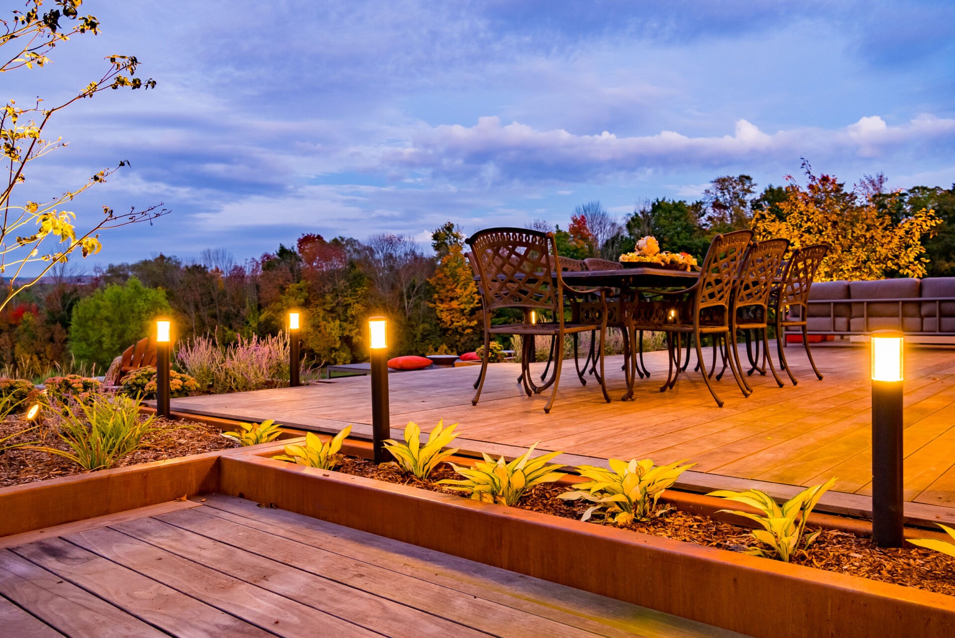 An inviting outdoor wooden deck features a dining set, warm lighting, and a backdrop of trees with autumnal colors at dusk.