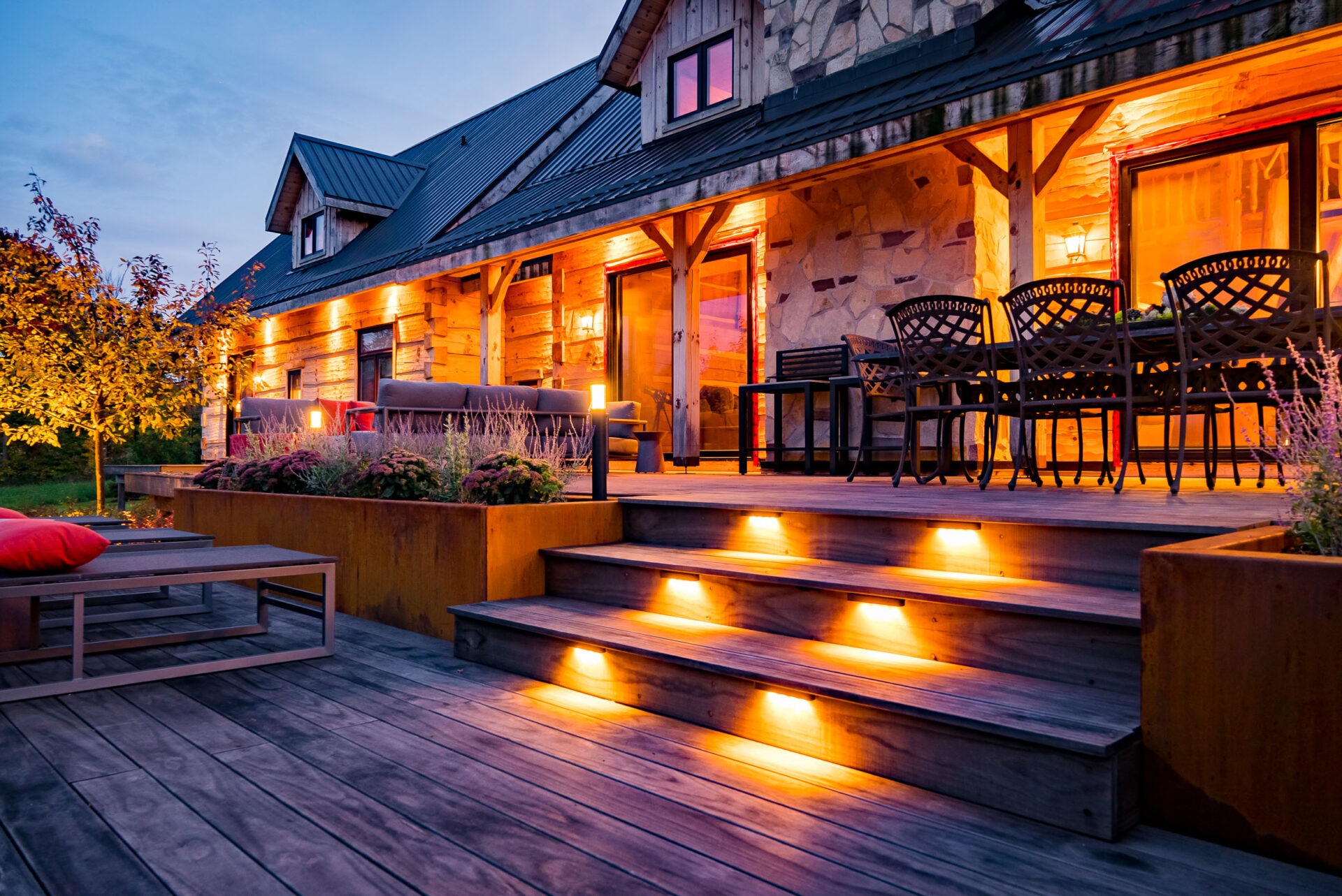A cozy wooden house with warm interior lighting. An inviting porch is adorned with outdoor furniture, steps with built-in lights, and potted plants. Twilight sets the mood.