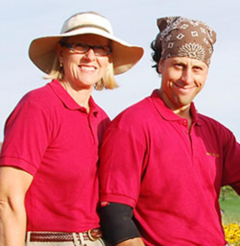 Two people stand side by side outdoors, wearing matching red polo shirts. One sports a wide-brimmed hat, and the other wears a bandana.
