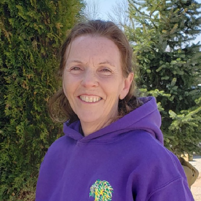 A smiling person in a purple hoodie stands outdoors, with a backdrop of green trees under a clear sky. They appear happy and relaxed.
