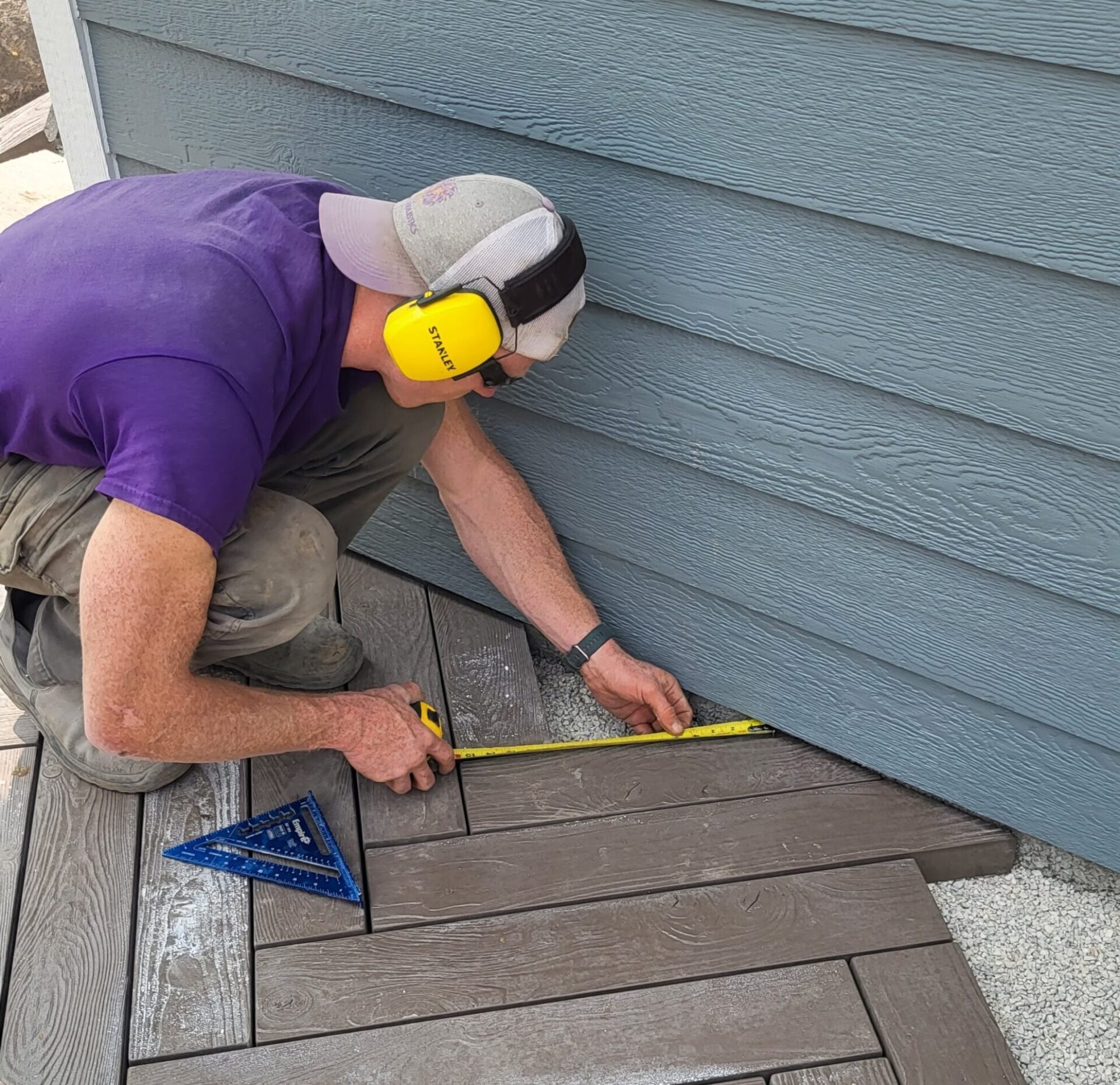 A person is kneeling on a deck, measuring with a tape measure, wearing a purple shirt, cap, and yellow ear protection. A speed square tool is beside them.