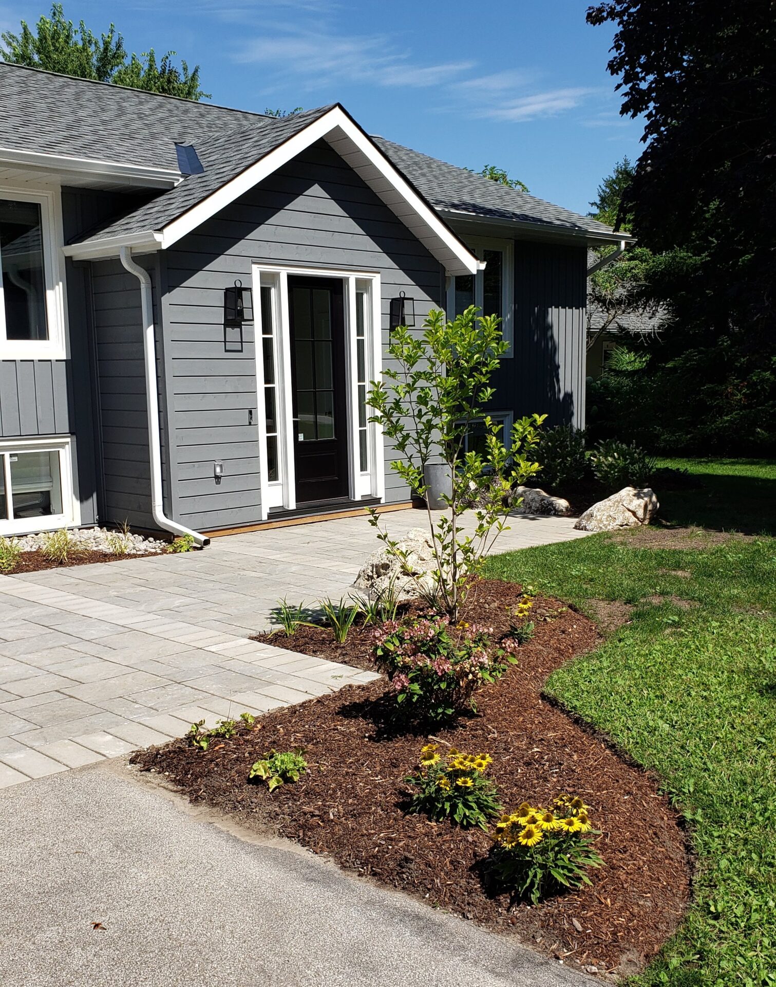 This is a residential grey house with a white-trimmed entryway, landscaping, a paver walkway, a small tree, flowering plants, and a sunny blue sky.