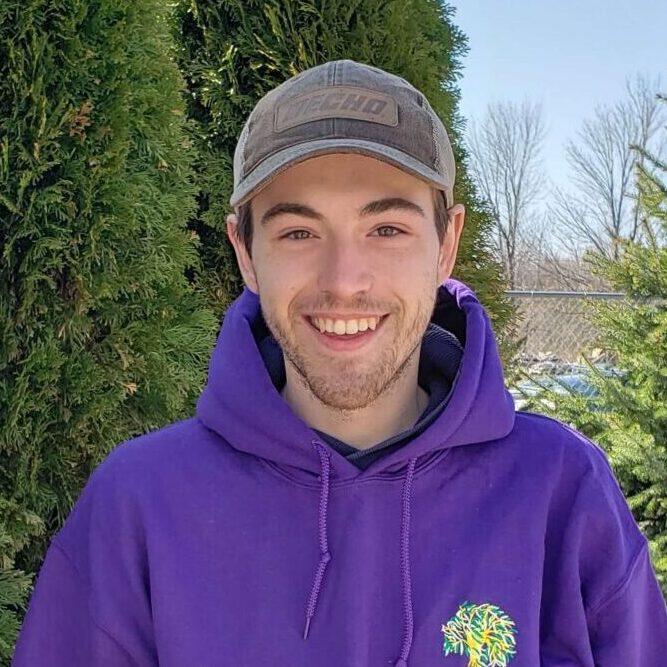 A person wearing a cap and a purple hoodie smiles in front of a green hedge under a clear blue sky. The person appears joyful and casual.