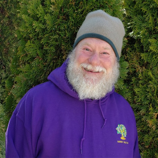 A smiling person with a white beard wearing a grey beanie and purple hoodie stands before a green bushy background.