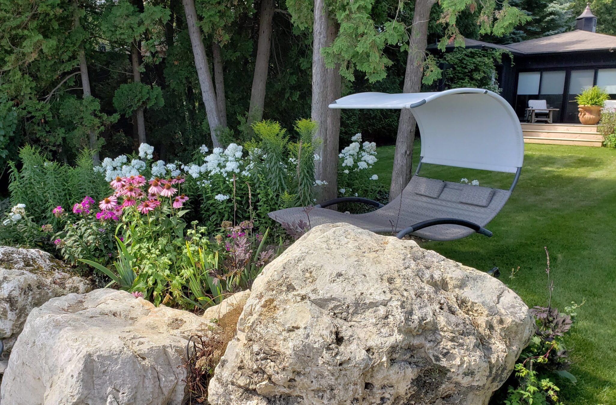A serene garden with vibrant flowers and a modern swing chair overlooking a well-manicured lawn, with a house in the background. Large rocks in foreground.