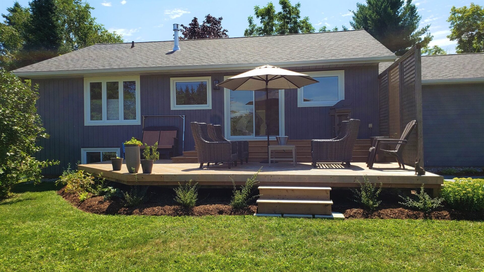 A backyard with a wooden deck featuring wicker furniture and an umbrella, adjacent to a dark gray house with white windows on a sunny day.