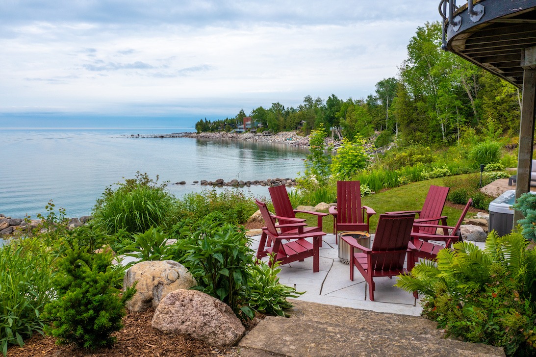 A serene lakeside setting with red Adirondack chairs, lush greenery, a clear sky, and a distant view of a rocky shoreline with a small building.