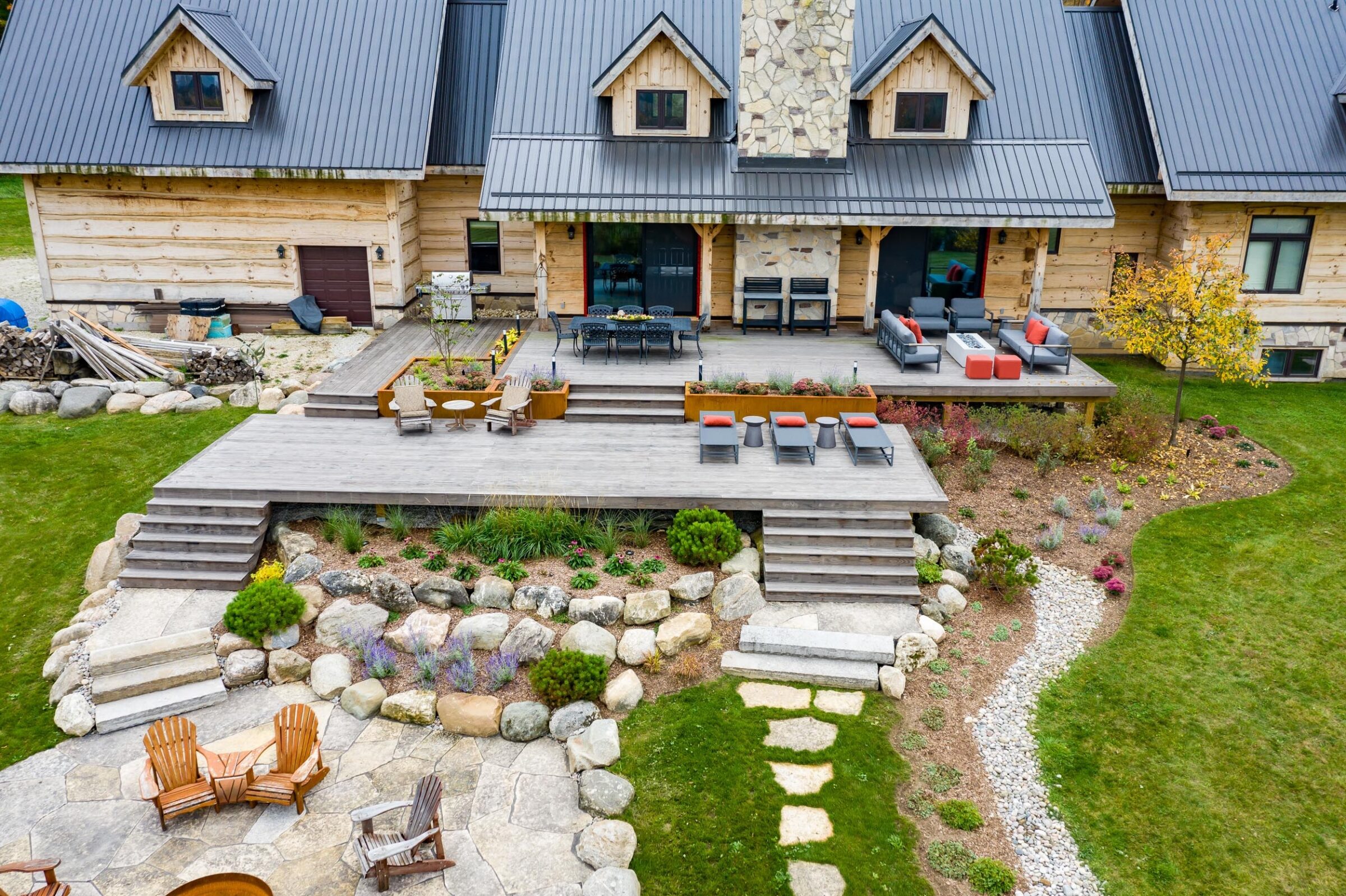 Aerial view of a stone and wood house with a terraced garden, outdoor seating areas, a lawn path, and a fire pit surrounded by Adirondack chairs.