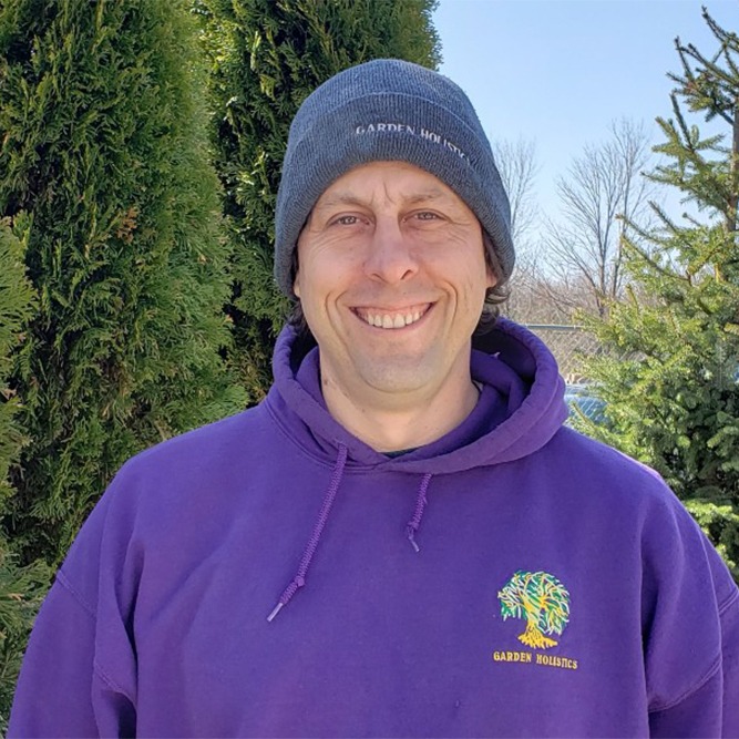 A person in a purple hoodie and grey beanie smiles against a backdrop of green coniferous trees on a sunny day.