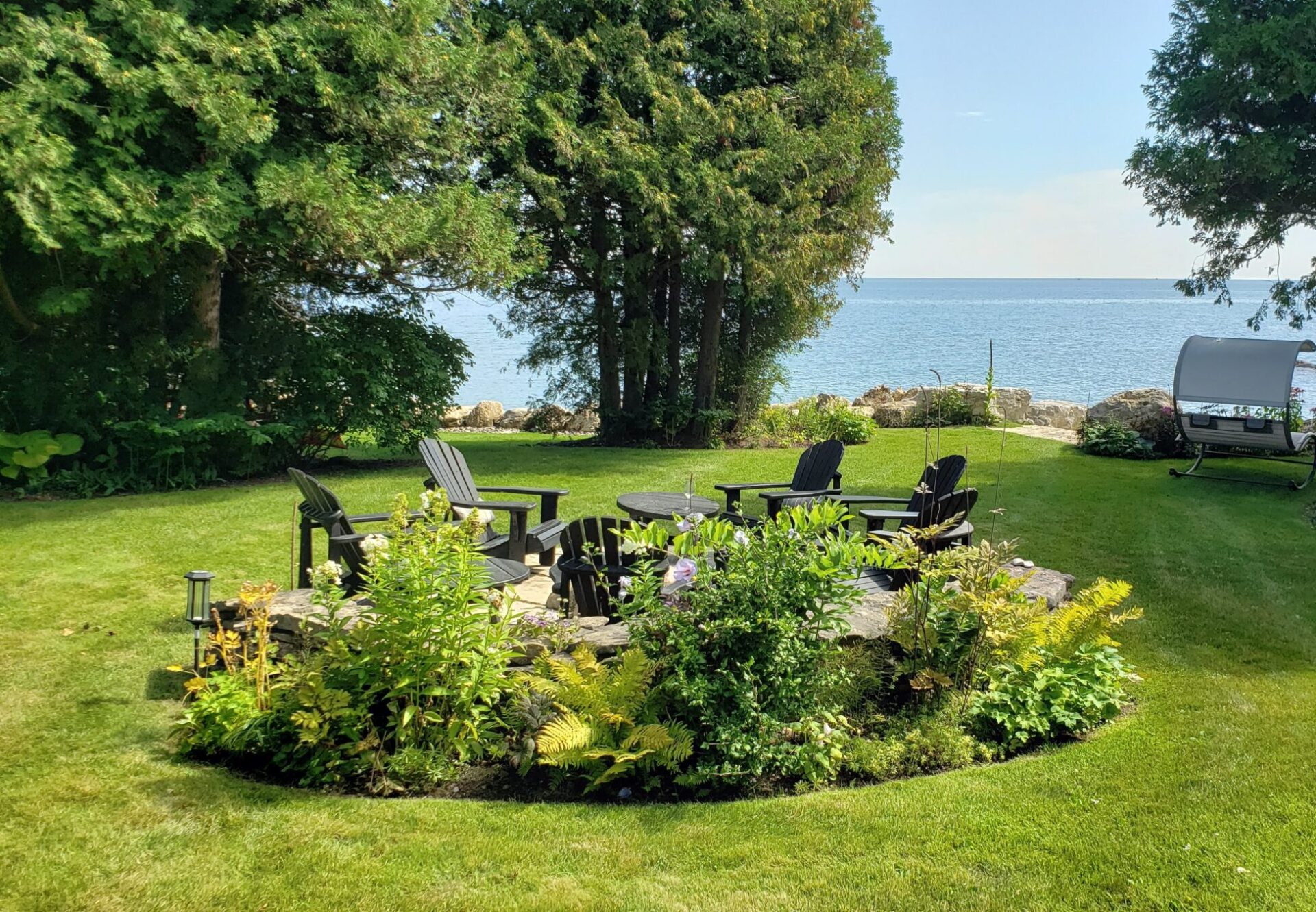 A serene garden with Adirondack chairs arranged around a fire pit, overlooking a calm sea, framed by lush greenery under a clear sky.