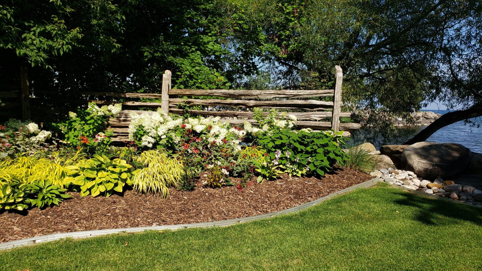 A lush garden with colorful flowers, a rustic wooden fence, and green grass, set against a backdrop of trees and a glimpse of water.