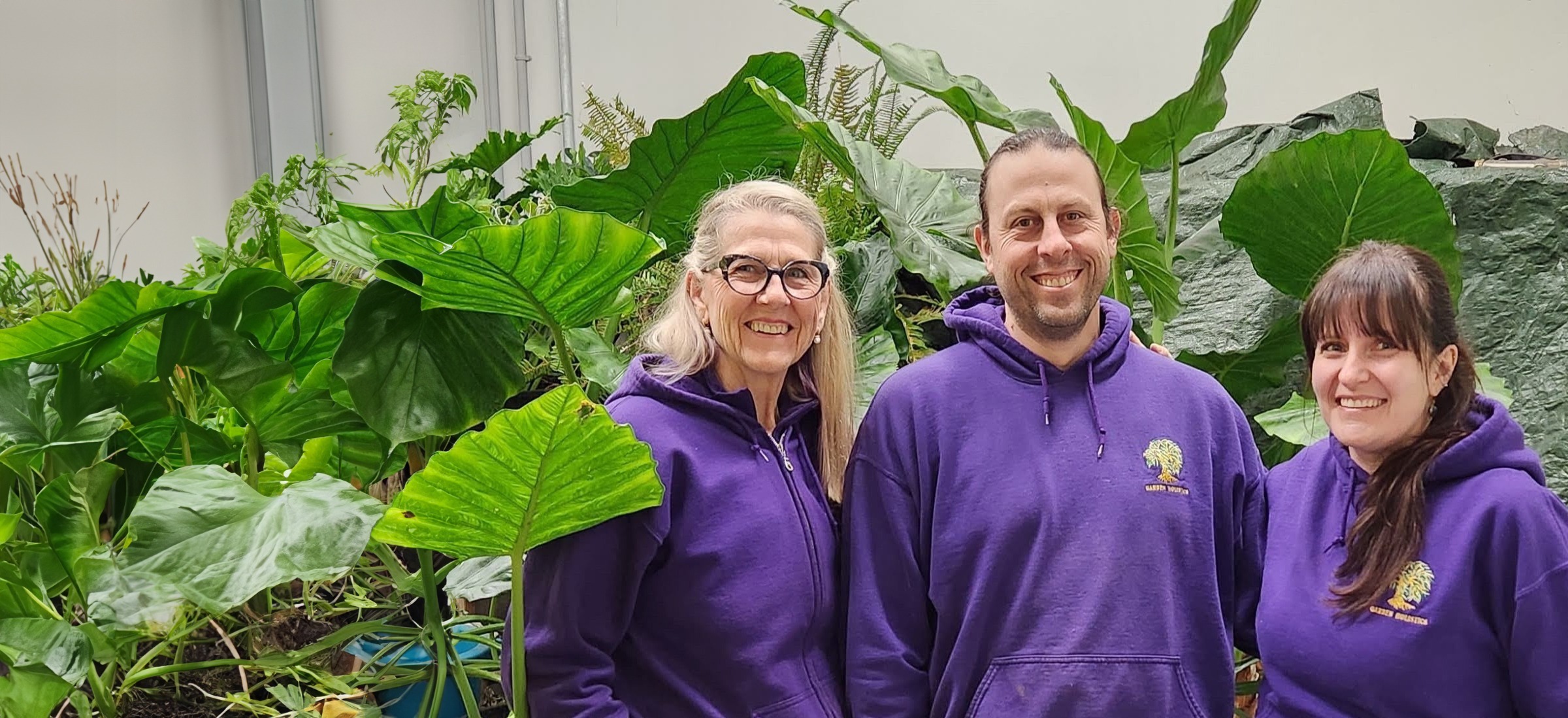 Three people stand in front of large green leaves, smiling and wearing matching purple hoodies with a logo, in a bright, plant-filled indoor setting.