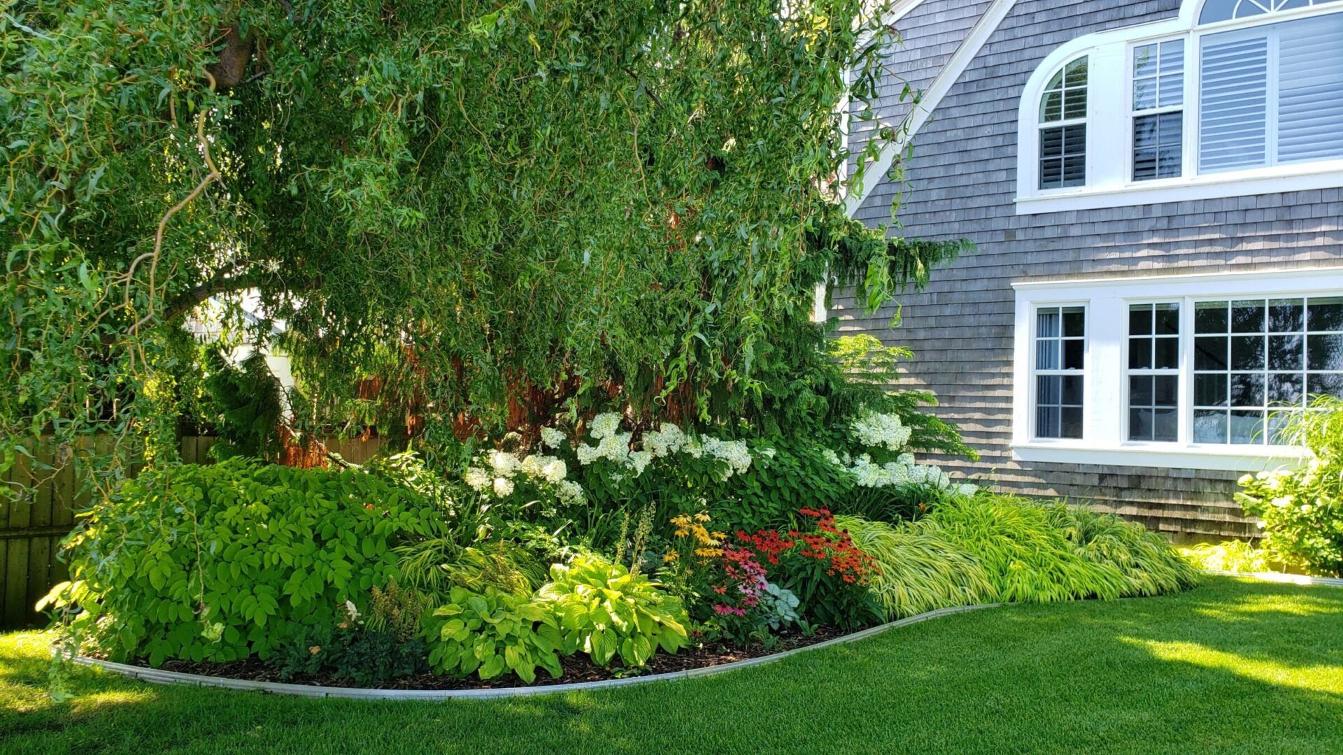 Lush garden with vibrant flowers next to a traditional house with white windows and gray shingles. The lawn is neatly trimmed and green.