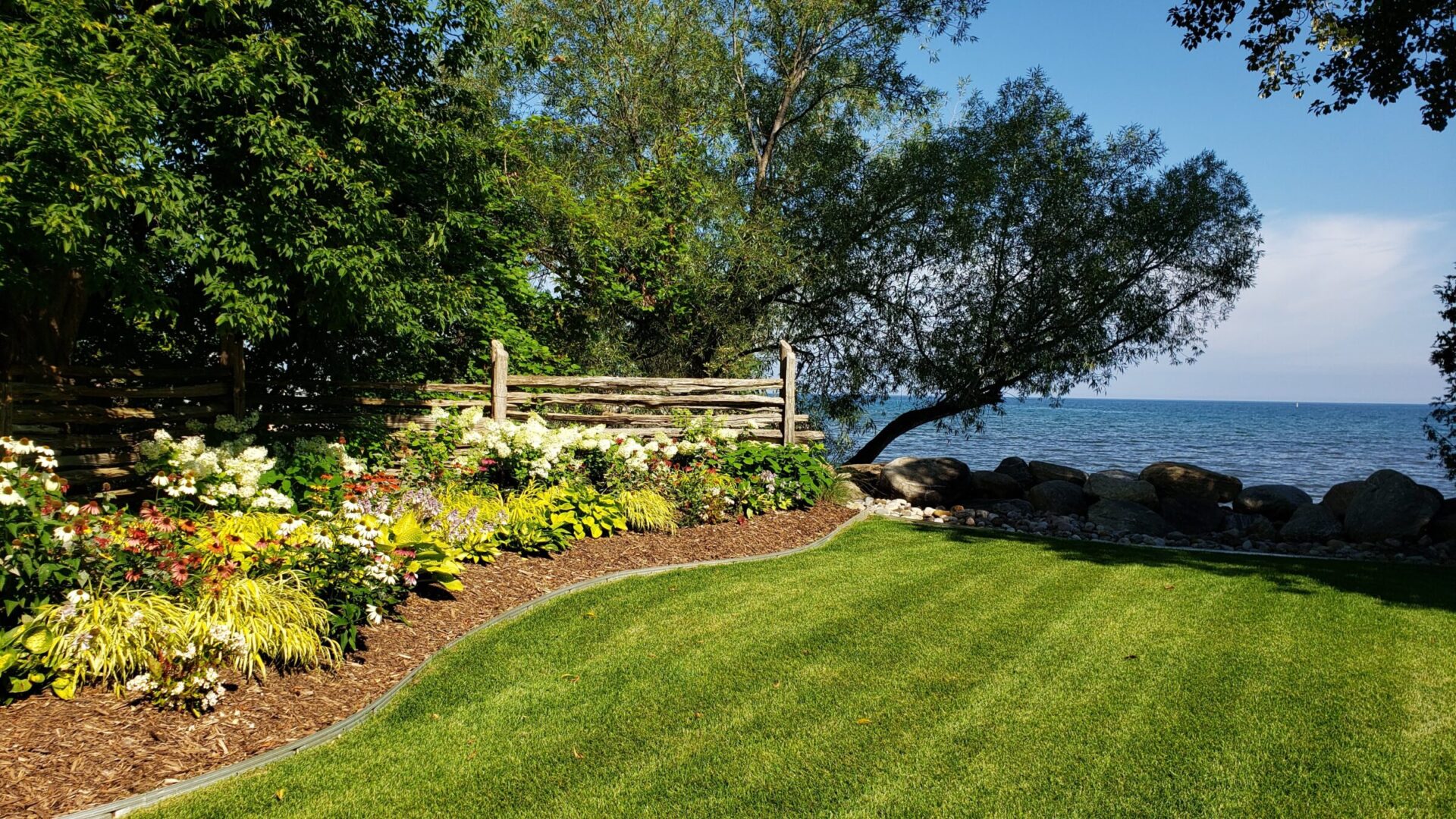 The image shows a serene lakefront garden with a wooden fence, lush green lawn, flowering plants, and a large body of water in the background.