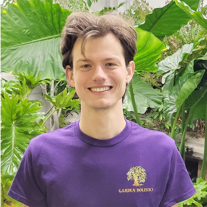 A smiling person with short hair stands in front of lush green leaves, wearing a purple t-shirt with a "Garden Holistics" logo.