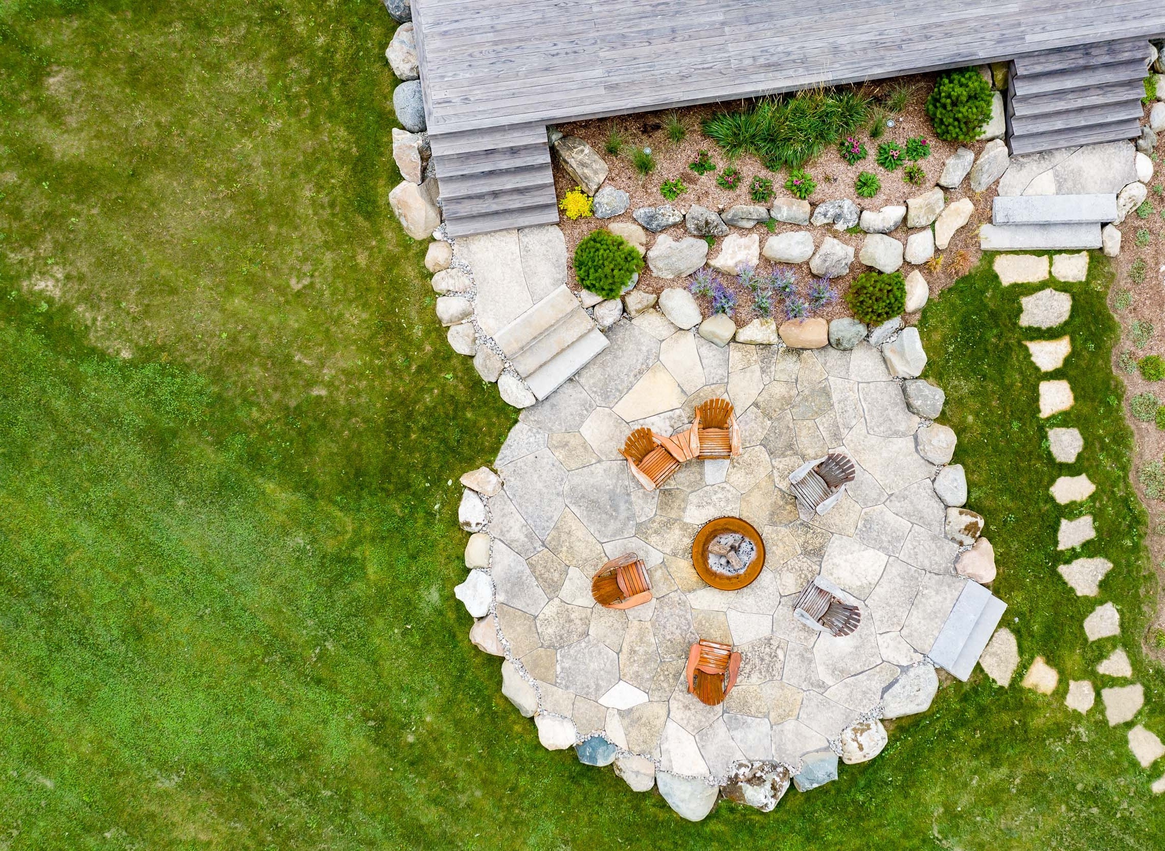 An aerial view of a landscaped backyard with a stone patio, fire pit, wooden chairs, a green lawn, stepping stones, and a grey wooden structure.