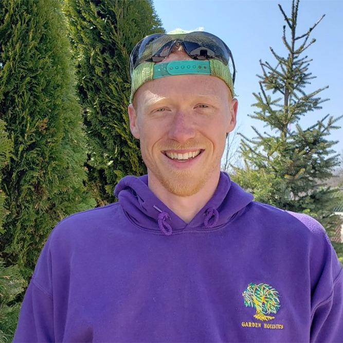 A smiling person wearing a purple hoodie and a cap stands before evergreen trees. The cap's visor reflects an image, possibly the photographer.