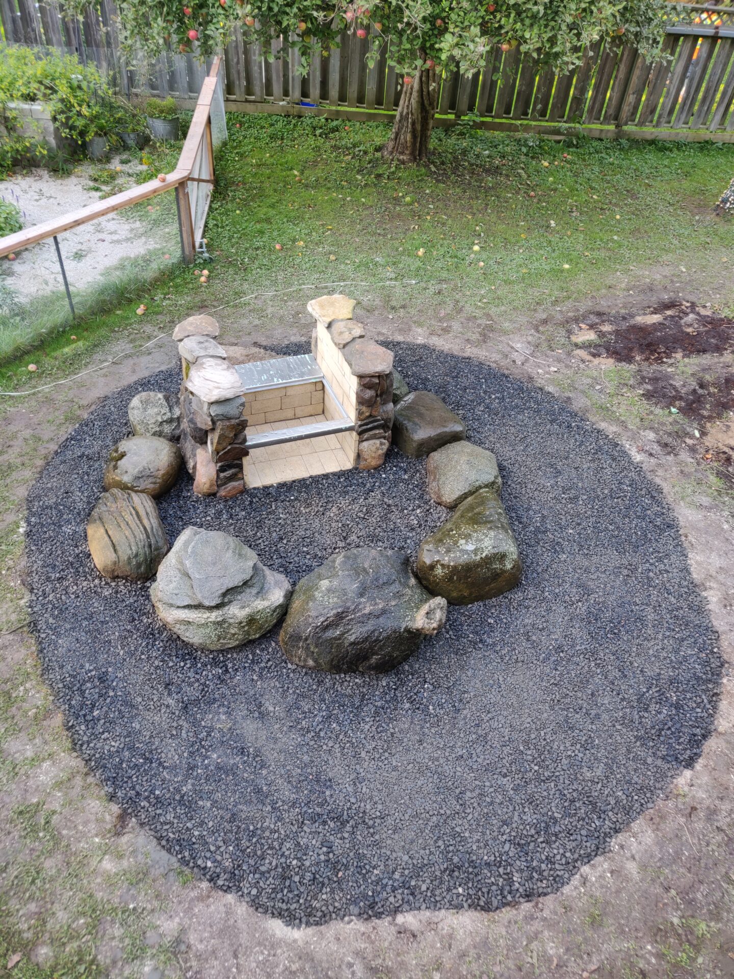 A backyard fire pit area with stone seating, surrounded by a circular gravel bed. Apples have fallen from a tree near a wooden fence.