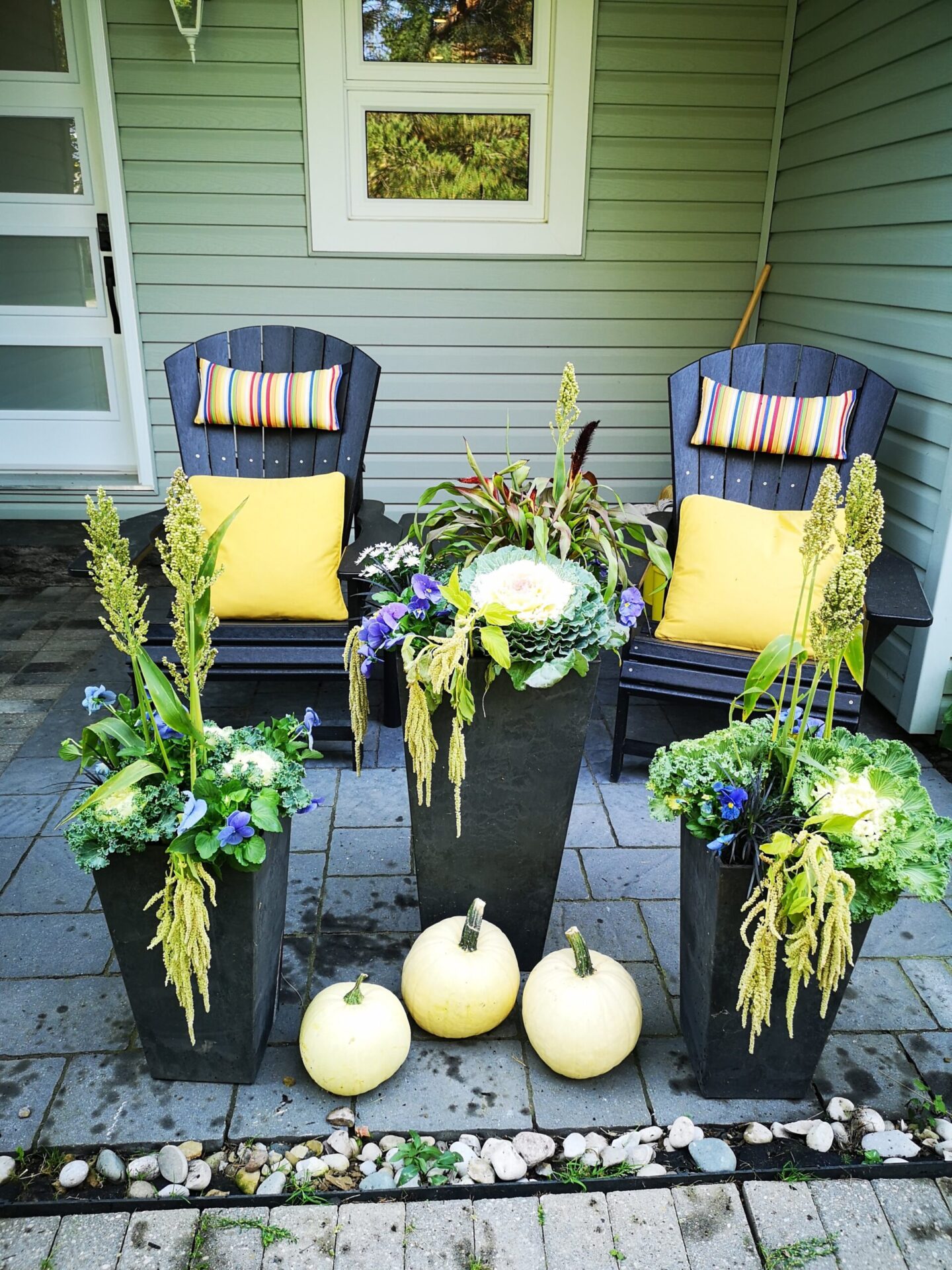 Two Adirondack chairs with colorful cushions sit on a patio, flanked by ornate planters and white pumpkins, with a green house background.