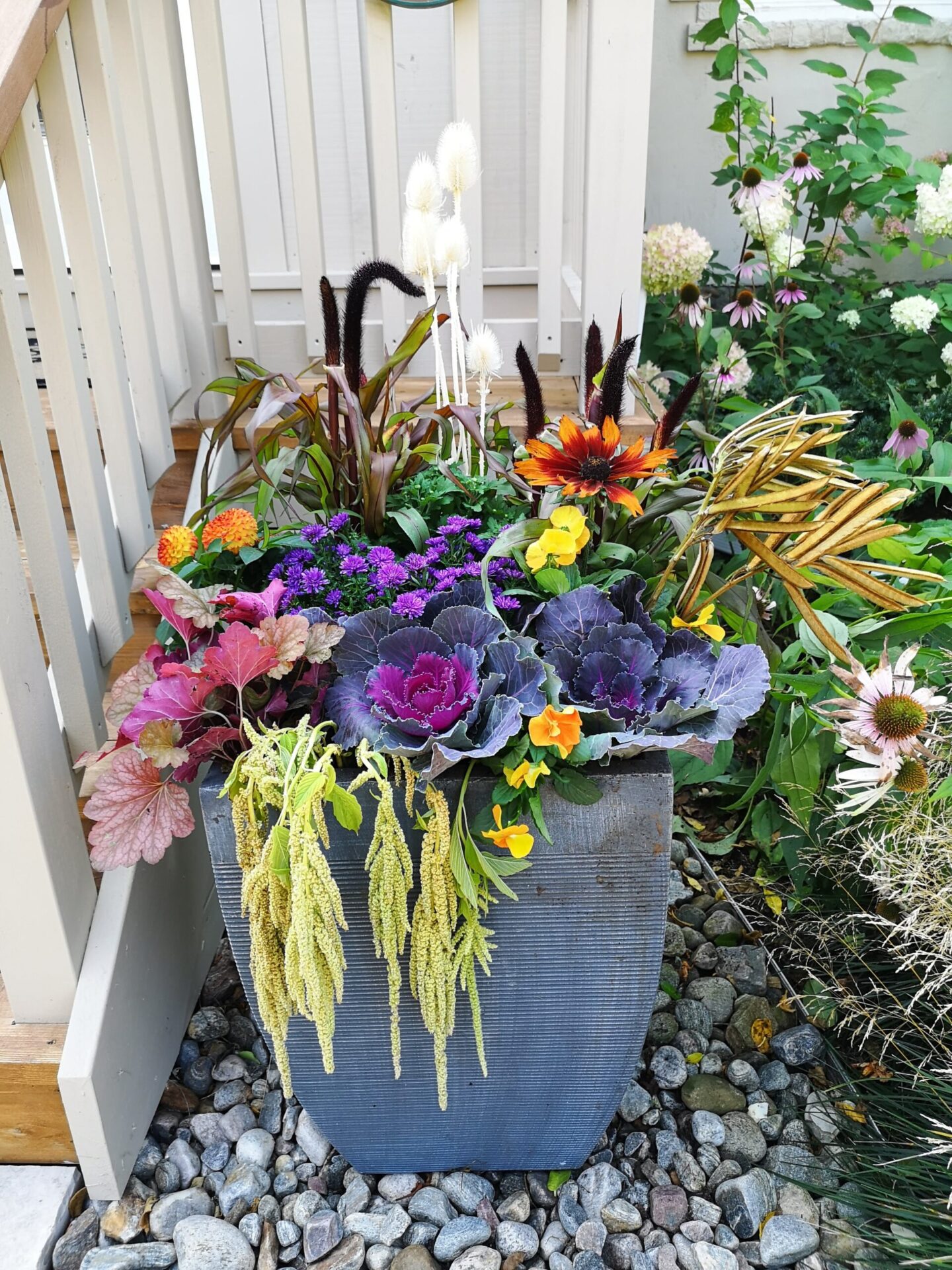 This image shows a vibrant arrangement of various flowers and plants in a large blue-gray pot, set against a pebble-covered ground and a wooden railing.