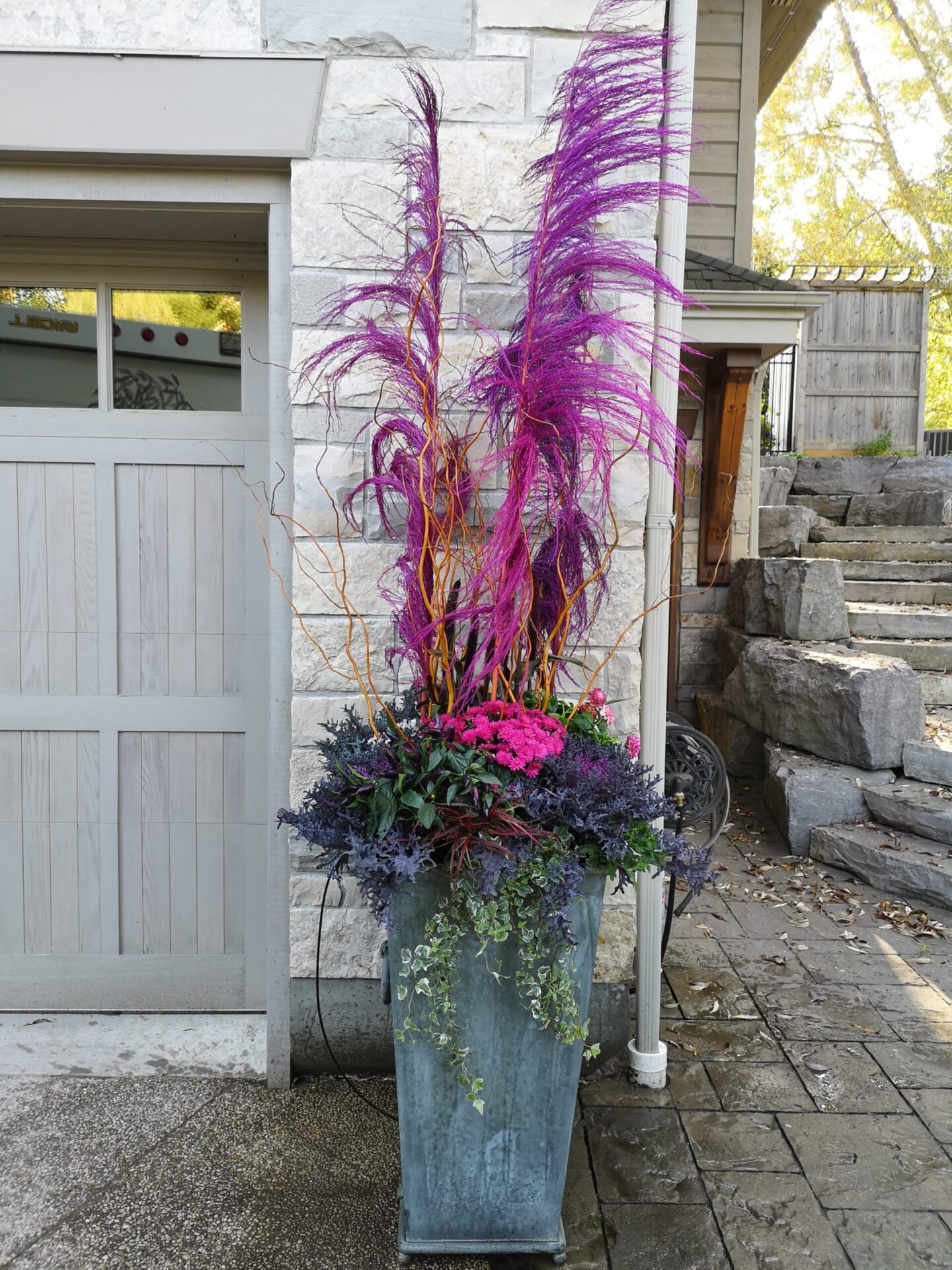 A large decorative planter with vibrant purple and pink flowers, feathery plumes, and trailing greenery placed beside a stone house entrance.