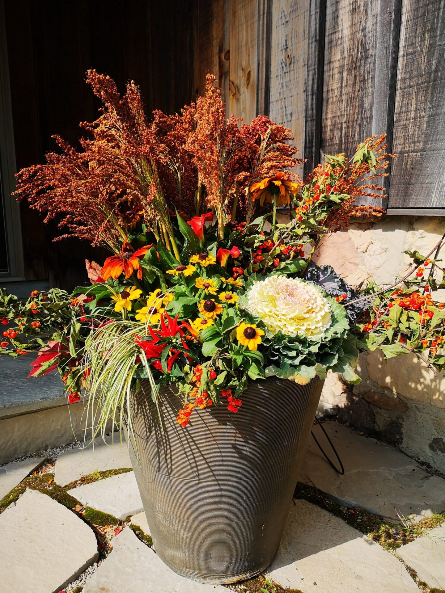 A large flower arrangement in a textured pot with red, yellow, and green plants, ornamental cabbage, and tall grasses, located on a paved surface by a wooden wall.