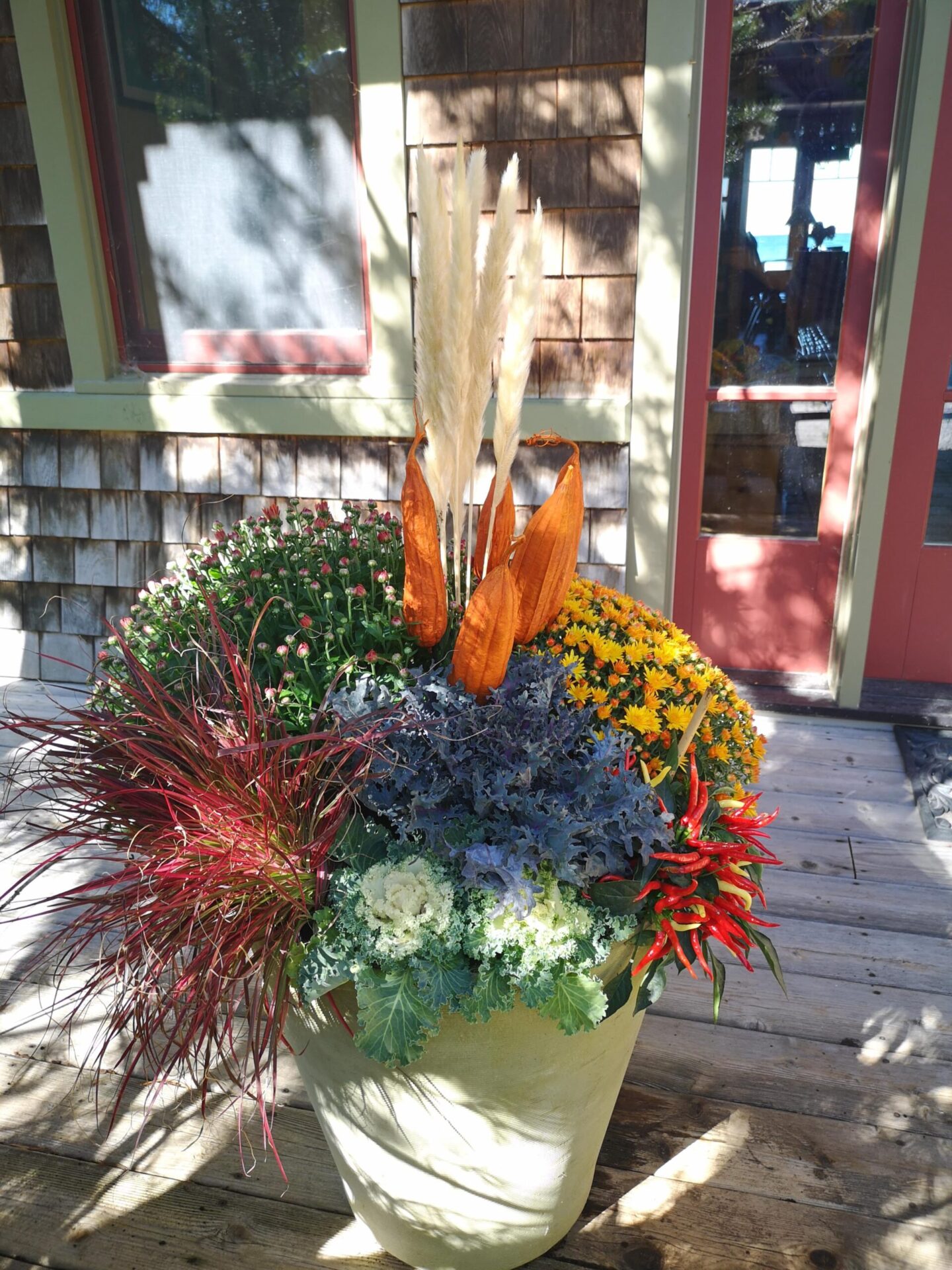 A vibrant decorative planter with various colorful plants and ornamental grasses on a wooden deck, against a house with cedar shingles and red trim.