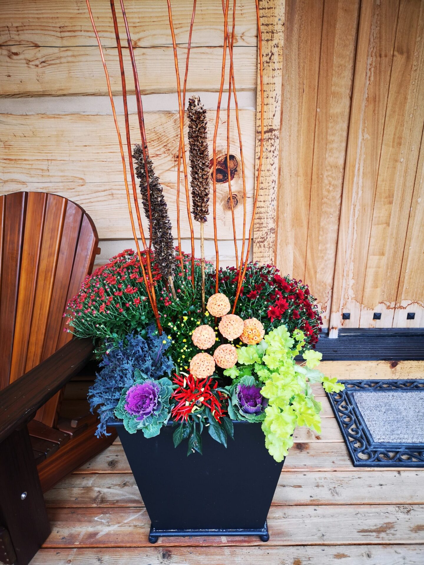 A vibrant floral arrangement in a black planter against a wooden background with tall decorative sticks, surrounded by rustic outdoor elements, including a bench.
