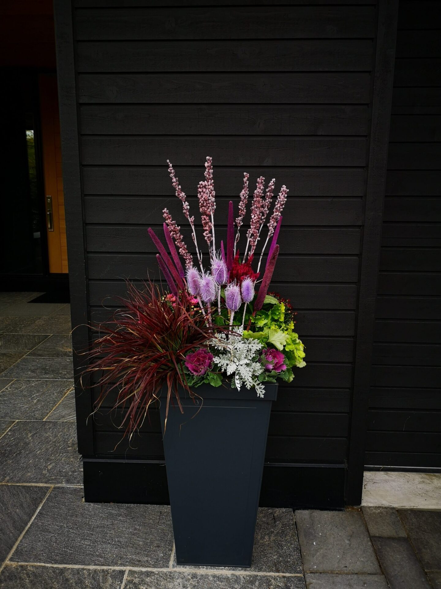 A tall black planter filled with vibrant flowers and ornamental grasses, set against a dark wooden backdrop by a stone paved area.