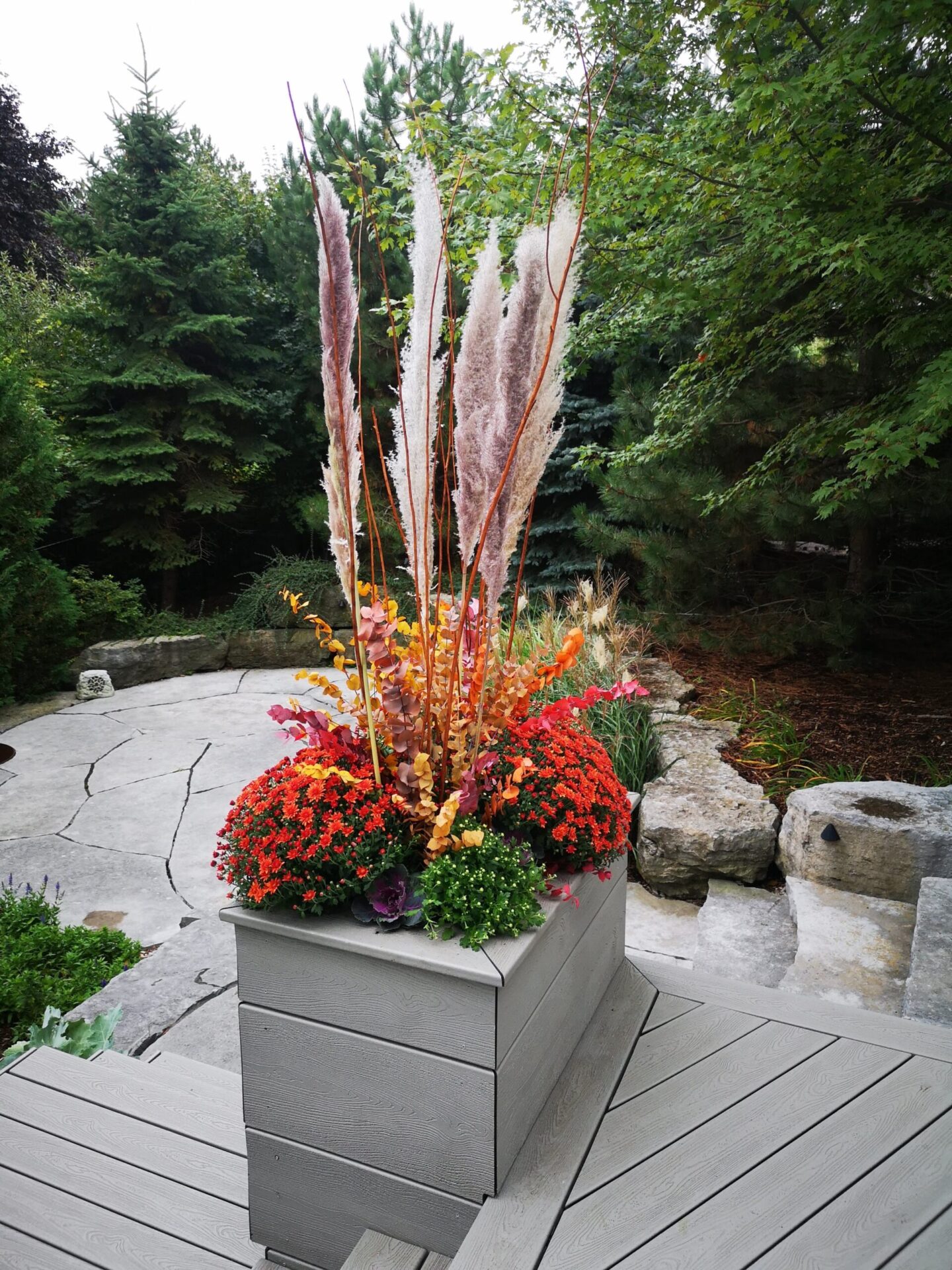This image features a vibrant floral arrangement in a gray planter on a wooden deck with tall trees and a stone pathway in the background.