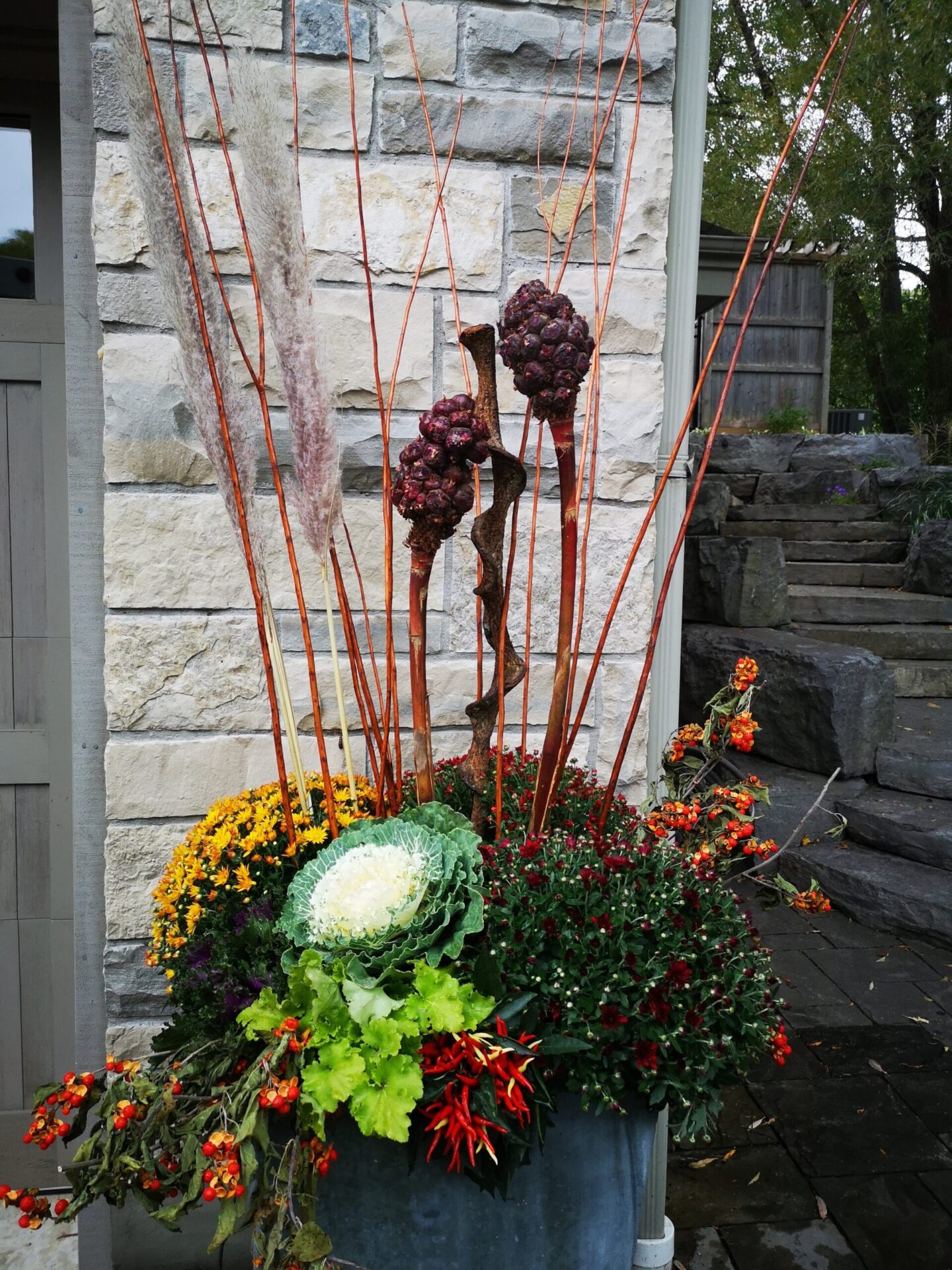 Outdoor autumn display with decorative cabbage, colorful flowers, and ornamental grasses in a large pot. Abstract sculptures resembling people with pinecone textures are behind.