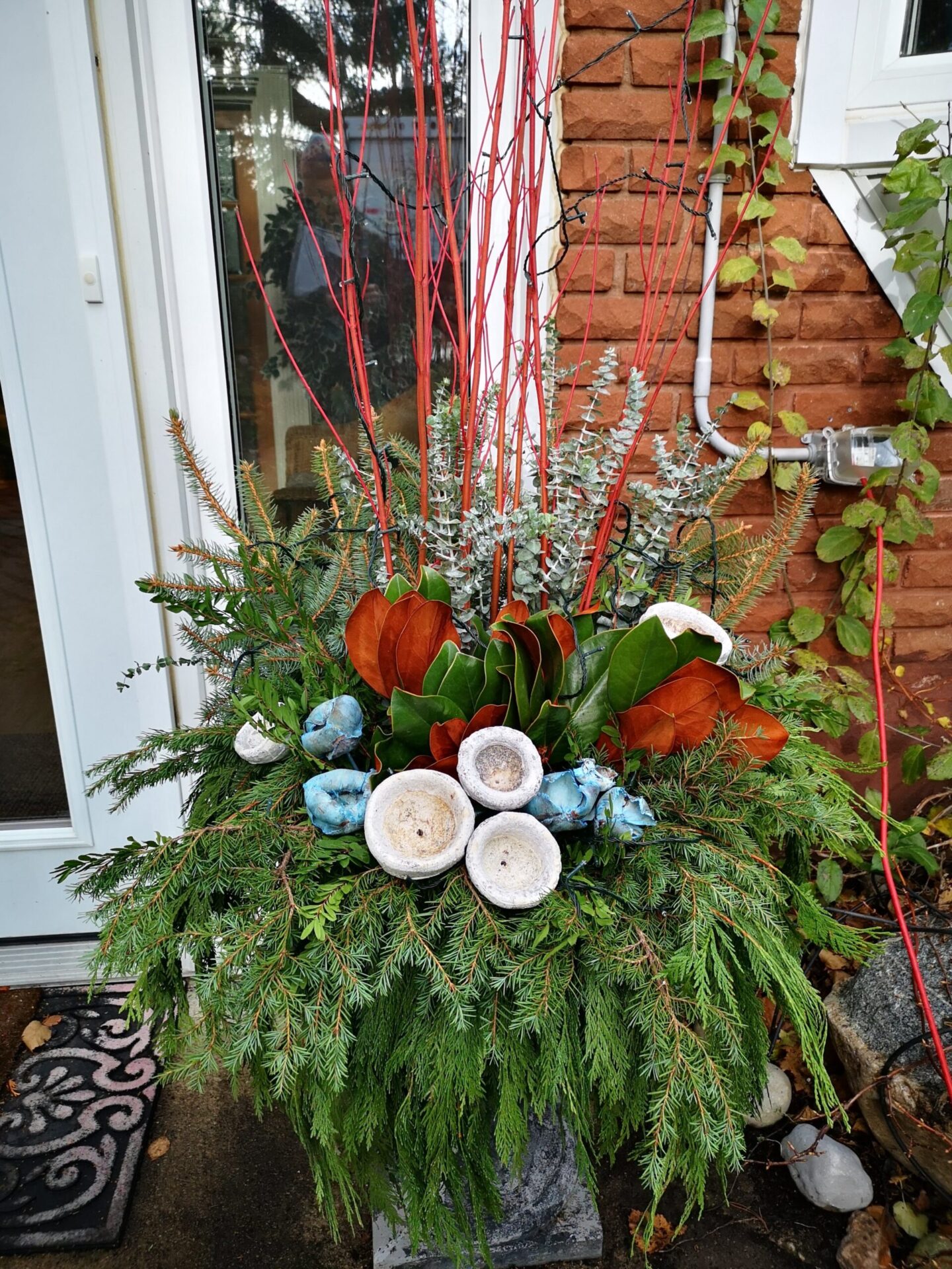 An outdoor floral arrangement featuring red branches, green foliage, blue ornaments, and white mushroom decorations in front of a house entrance.