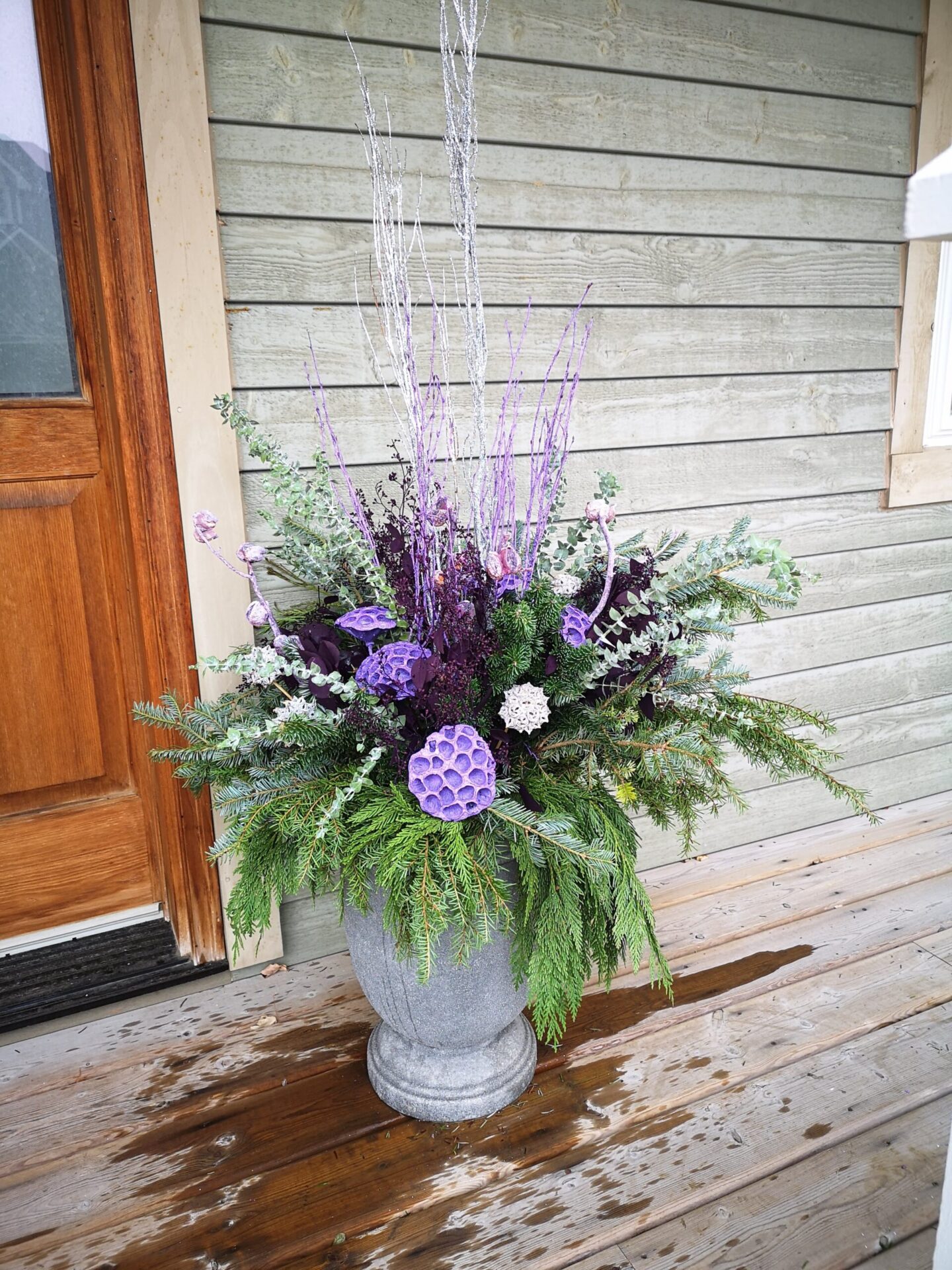 A decorative flower arrangement in a gray vase on a wet wooden porch, featuring purple flowers and greenery, with a wooden wall and door nearby.