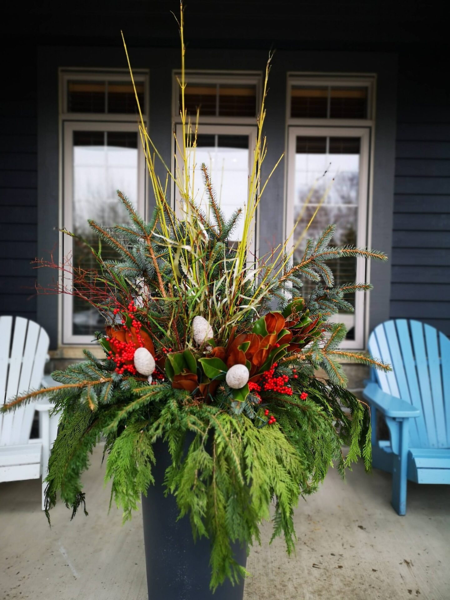 A vibrant floral arrangement in a planter with red berries, green foliage, and white accents, set against a porch with blue chairs and a dark house.