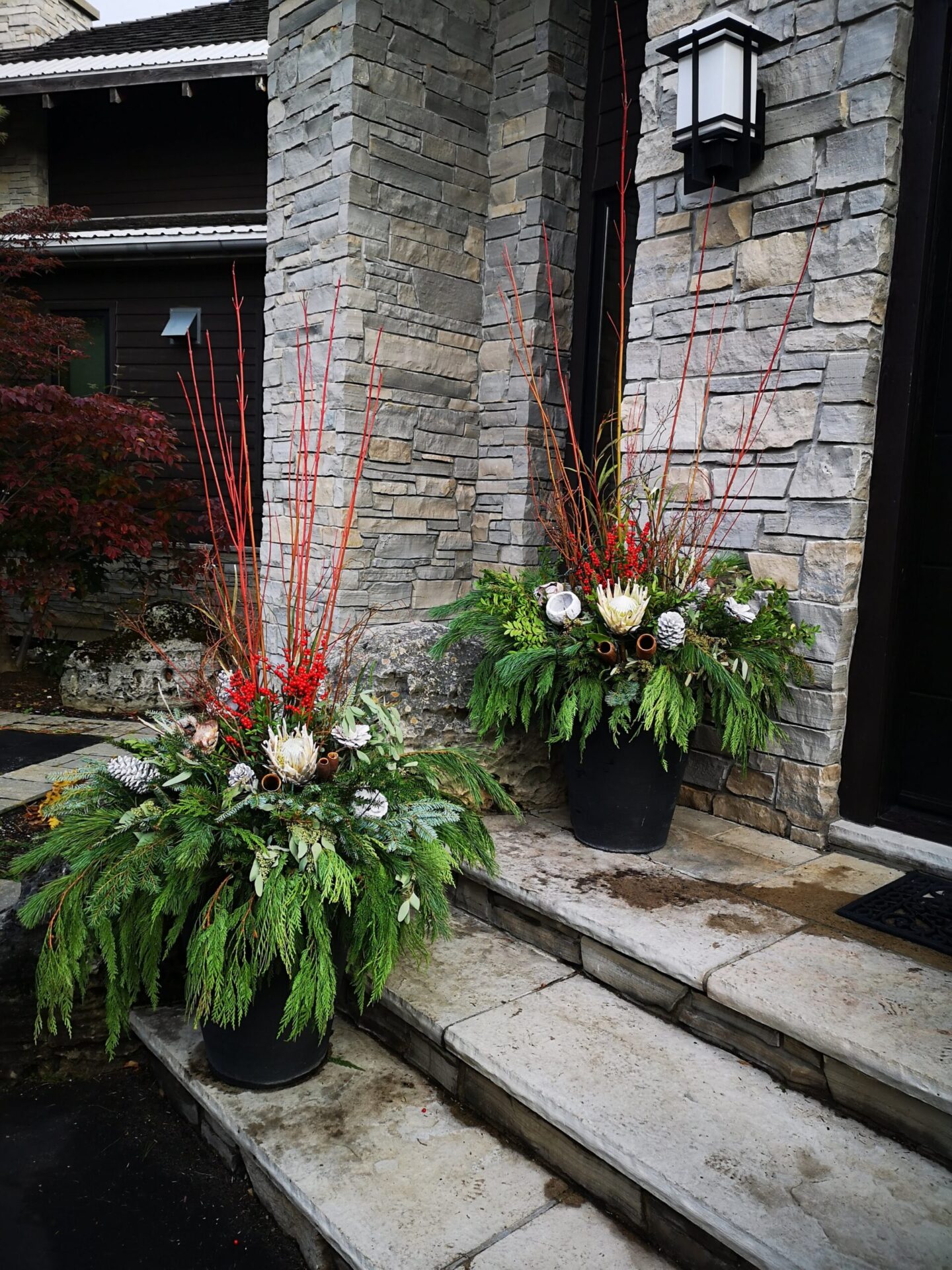 Two large floral arrangements in black pots stand on a stone stairway leading to a house with gray stone walls and a dark door.